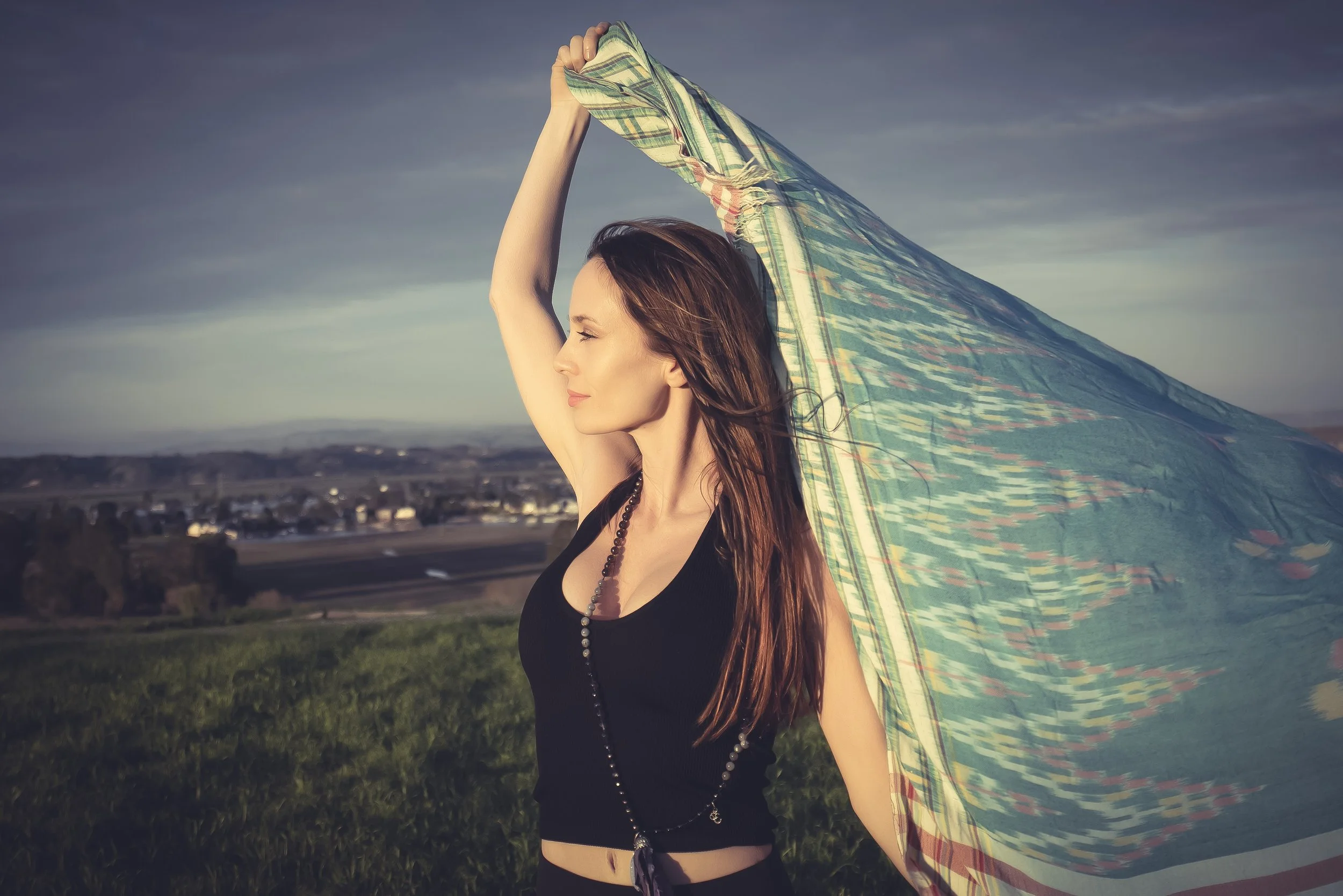 A woman with long hair is outdoors, holding a colorful scarf above her head, with her eyes closed and a serene expression, against a scenic landscape with a cloudy sky.