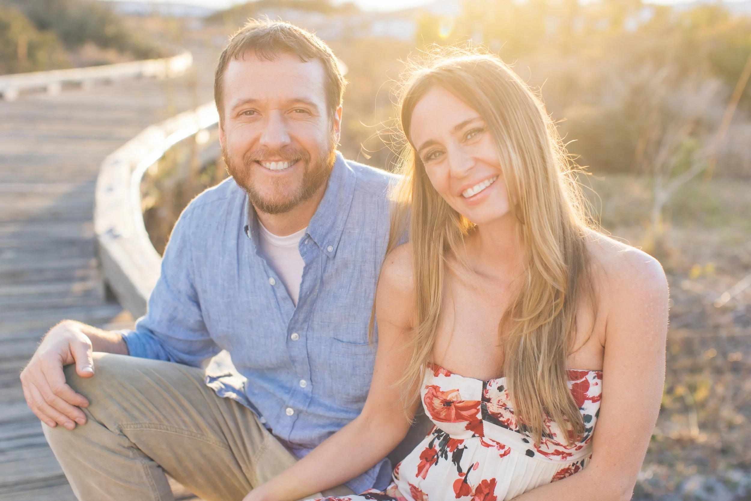 A smiling man and woman sitting together on a wooden bridge outdoors with sunlight shining behind them, in a natural setting with trees in the background.
