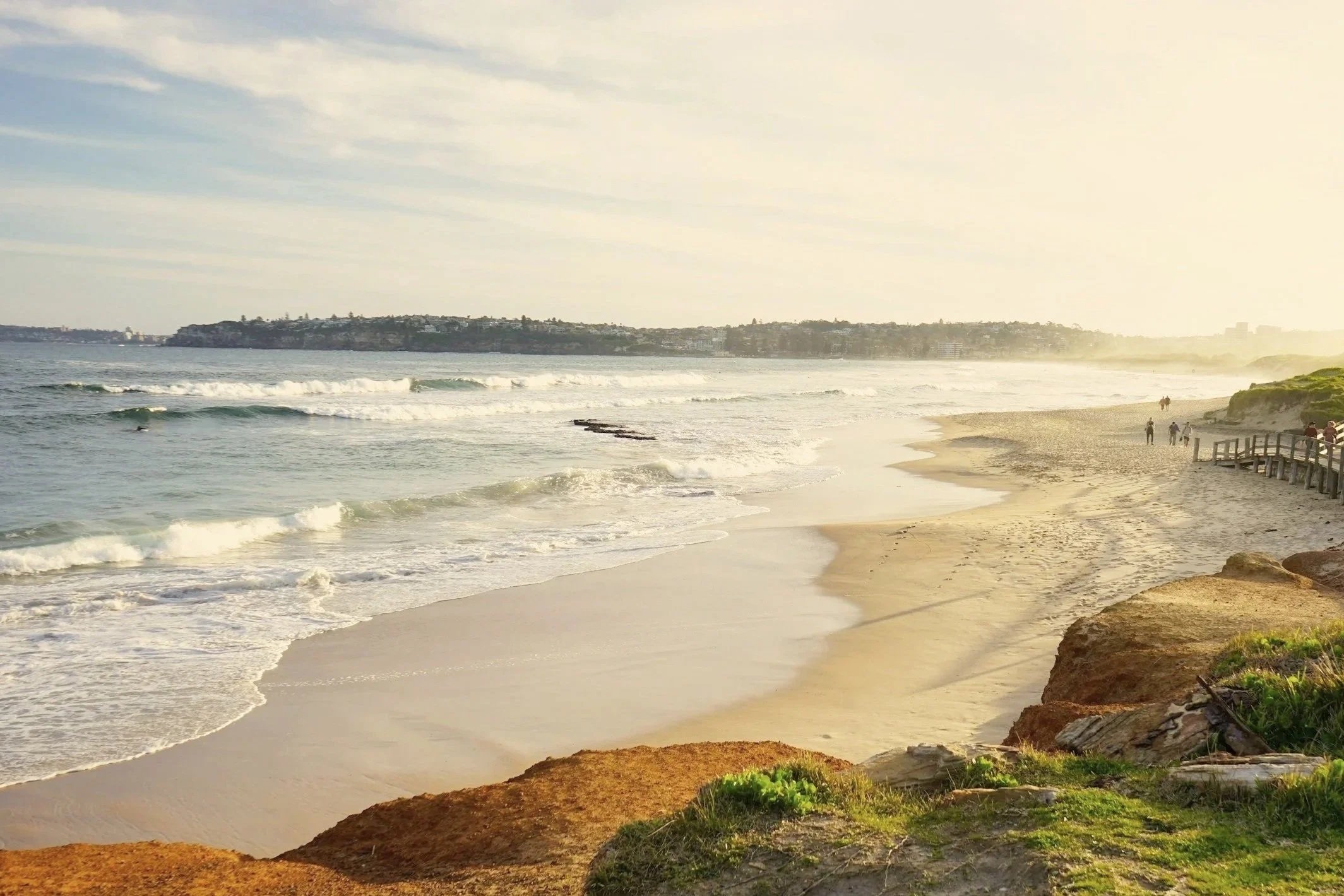 Beach scene with waves, sandy shore, rocks, green vegetation, and a few people walking along the beach during sunset or sunrise.