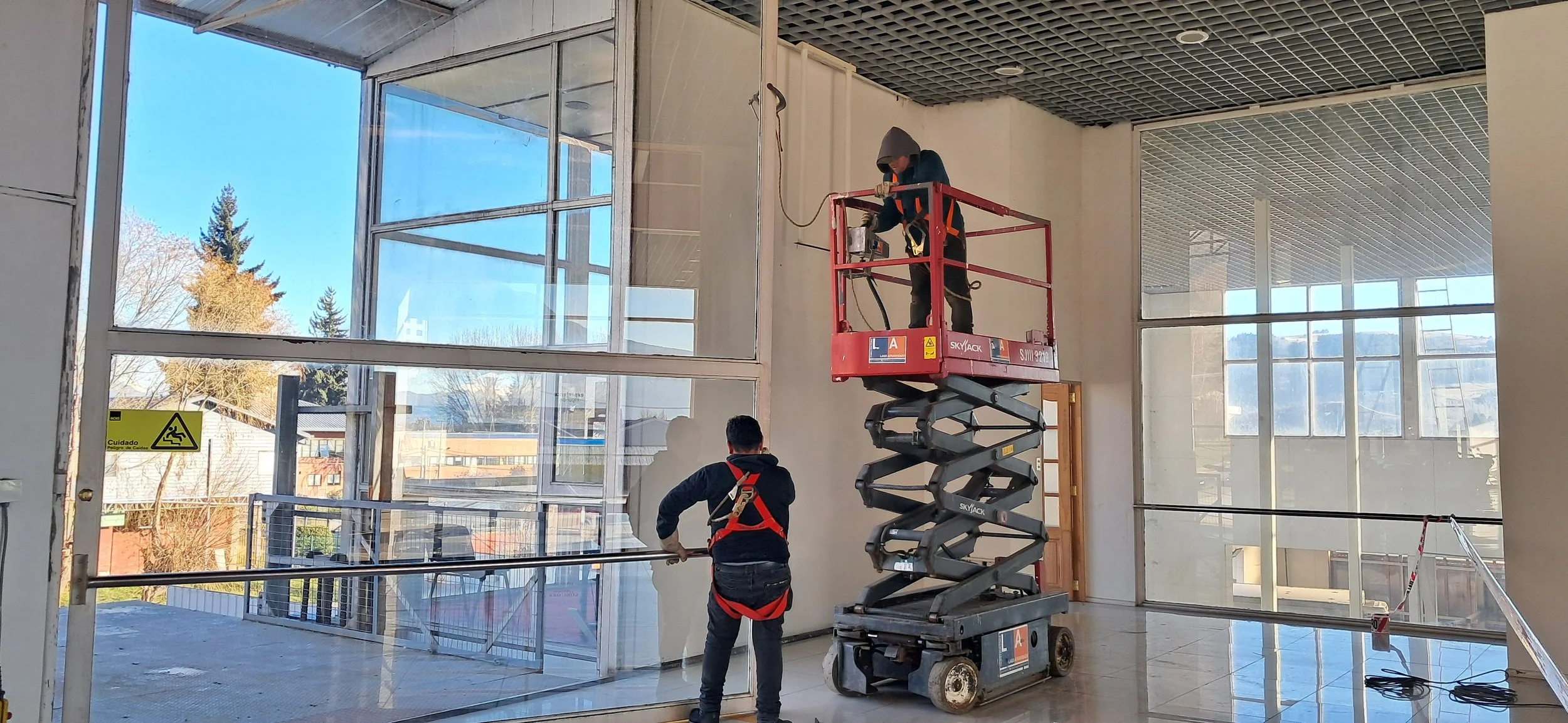 Dos trabajadores en elevador de tijera instalando grandes ventanas de vidrio en un edificio en construcción o renovación. La escena muestra un interior con paredes blancas, un techo de rejilla y muchas ventanas grandes con vista al exterior, incluyendo árboles y estructuras.