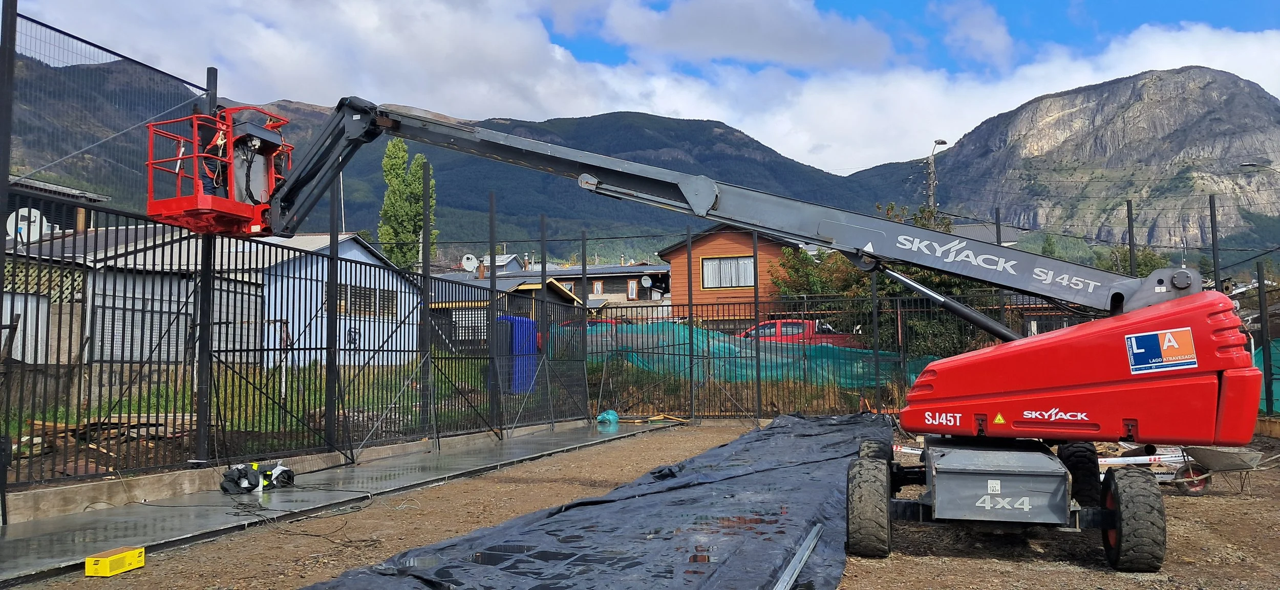 Maquinaria de construcción, una plataforma elevadora y una grúa en un área al aire libre con montaña y cielo nublado de fondo.