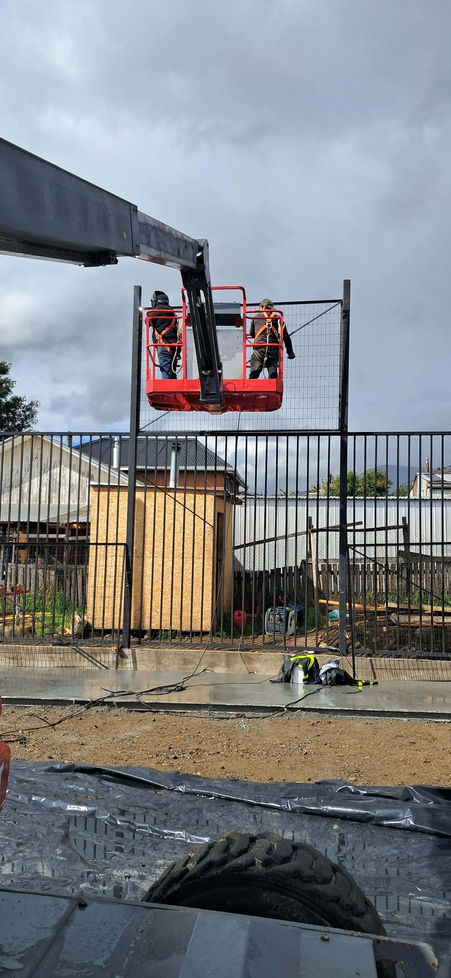 Dos trabajadores en una plataforma elevadora están instalando una cerca metálica en un área de construcción residencial con casas en el fondo. El cielo está nublado y parece que está por llover.