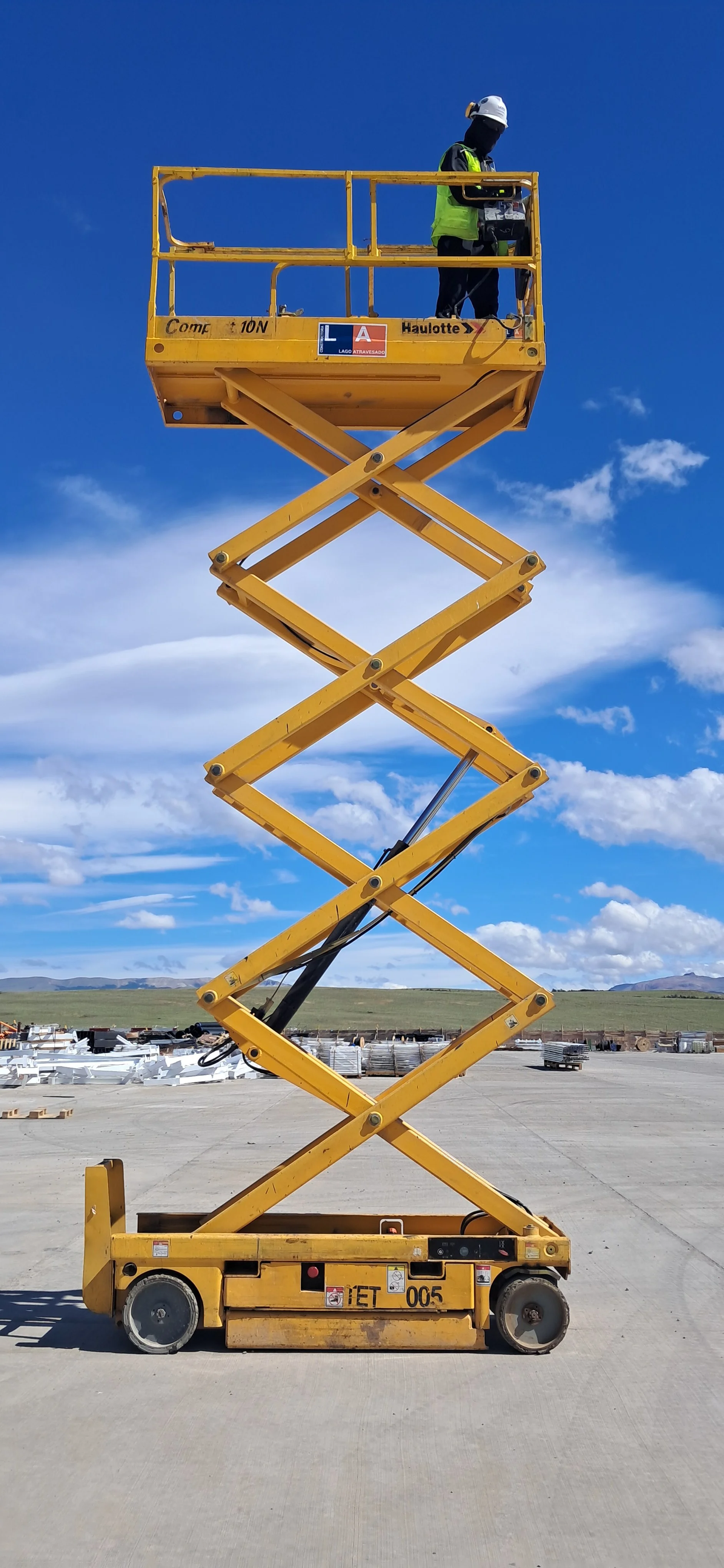 Persona en plataforma elevadora amarilla en un espacio abierto con cielo azul y nubes en el fondo.
