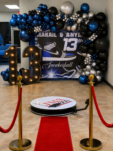 Balloon arch with black, blue, silver, and gold balloons framing a banner for a 13th birthday celebration, with large illuminated number 13, a red carpet, and a sneaker-themed backdrop. The event appears to be for someone named Makail and Anly, celeb