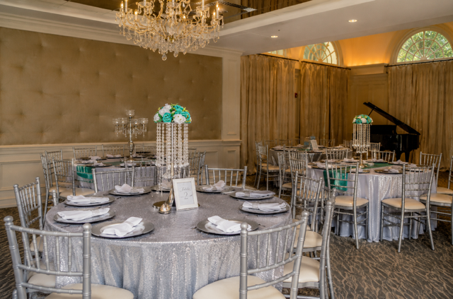 Elegant banquet room with round tables covered in silver tablecloths, decorated with floral centerpieces, silverware, and glassware, with a grand chandelier overhead.