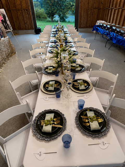 Long banquet table decorated for a formal event with white tablecloths, blue and gold dinnerware, napkins, glasses, and flowers, set inside a barn with sliding doors open to a green outdoor area.