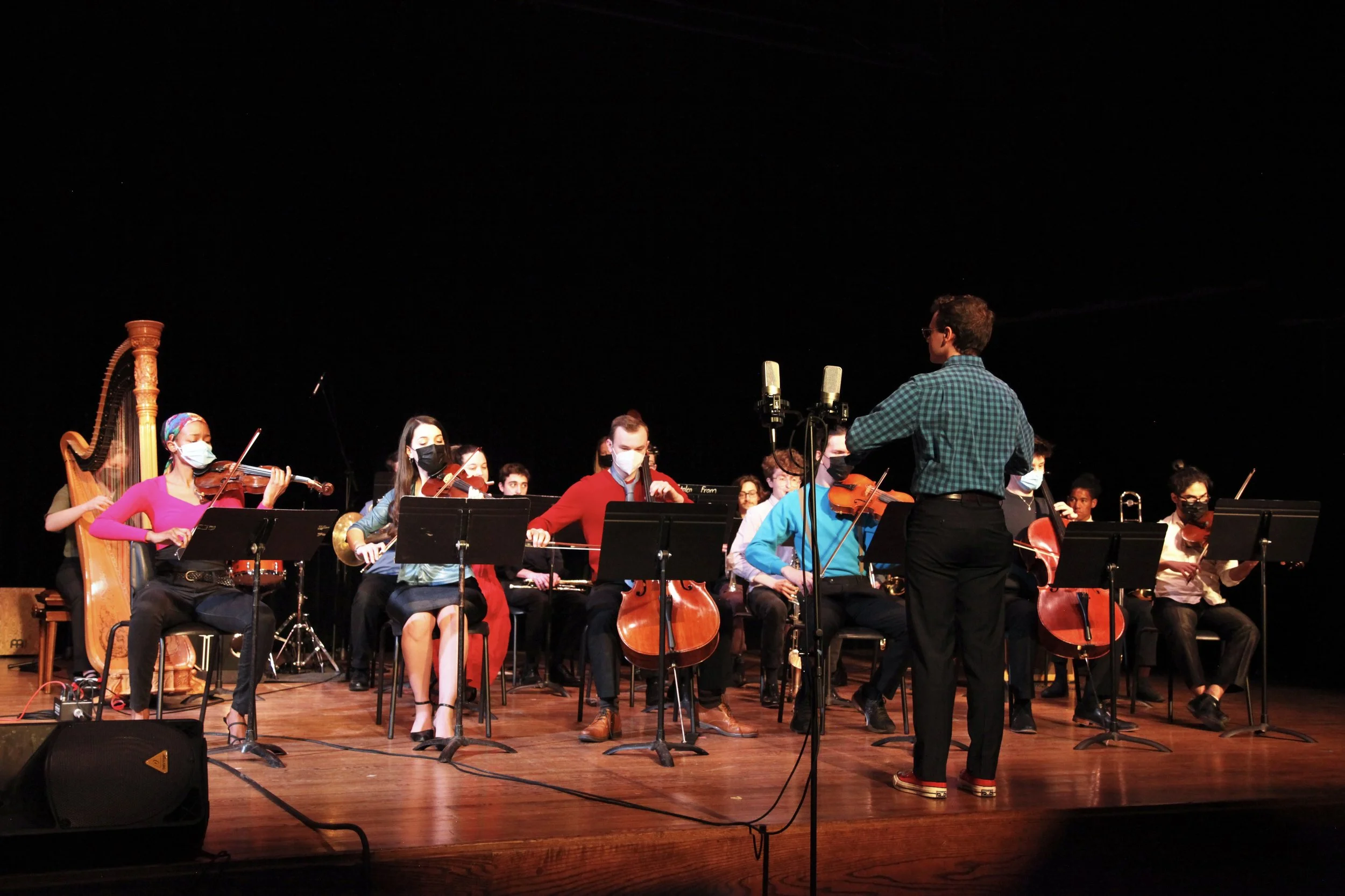 An orchestra performing on stage with musicians wearing face masks, led by a conductor standing in front of them. The stage has microphones and music stands, and the backdrop is black.