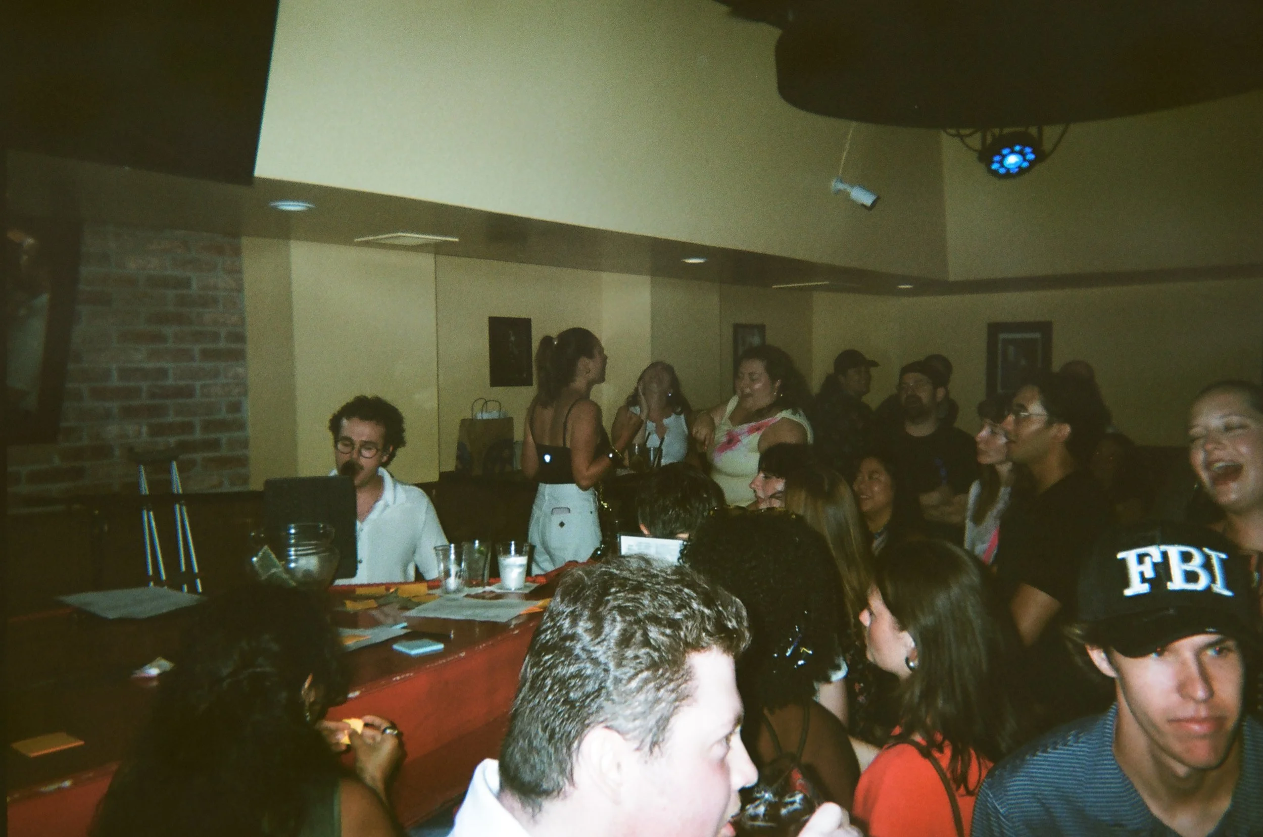 A group of people gathered at Georgetown Piano Bar, some are engaged in conversation while others are sitting or standing. There is a red table with papers and cups in the foreground, and Andrew is performing in the background.