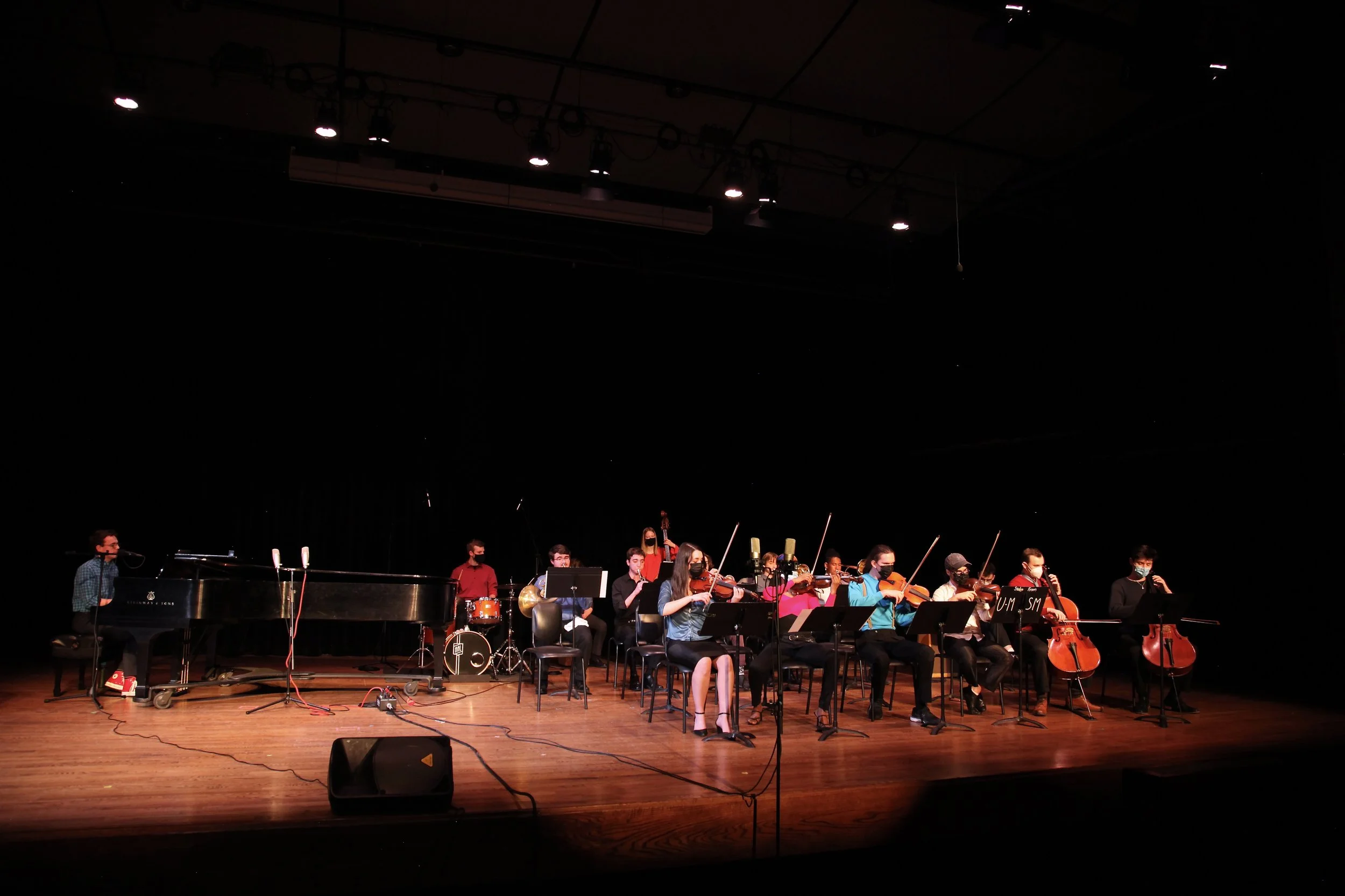 A group of musicians on a stage, including a pianist, playing various instruments such as violins, cello, and drums, in a concert hall with a black backdrop and stage lighting.
