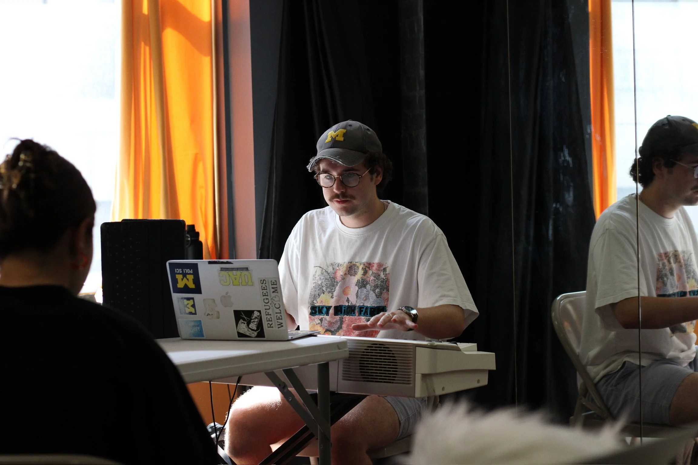A man with curly hair, glasses, and a mustache sits at a table with a laptop covered in stickers, including ones with the University of Michigan and other designs. He is wearing a gray cap with a yellow 'M' logo and a white T-shirt with graphics. In 