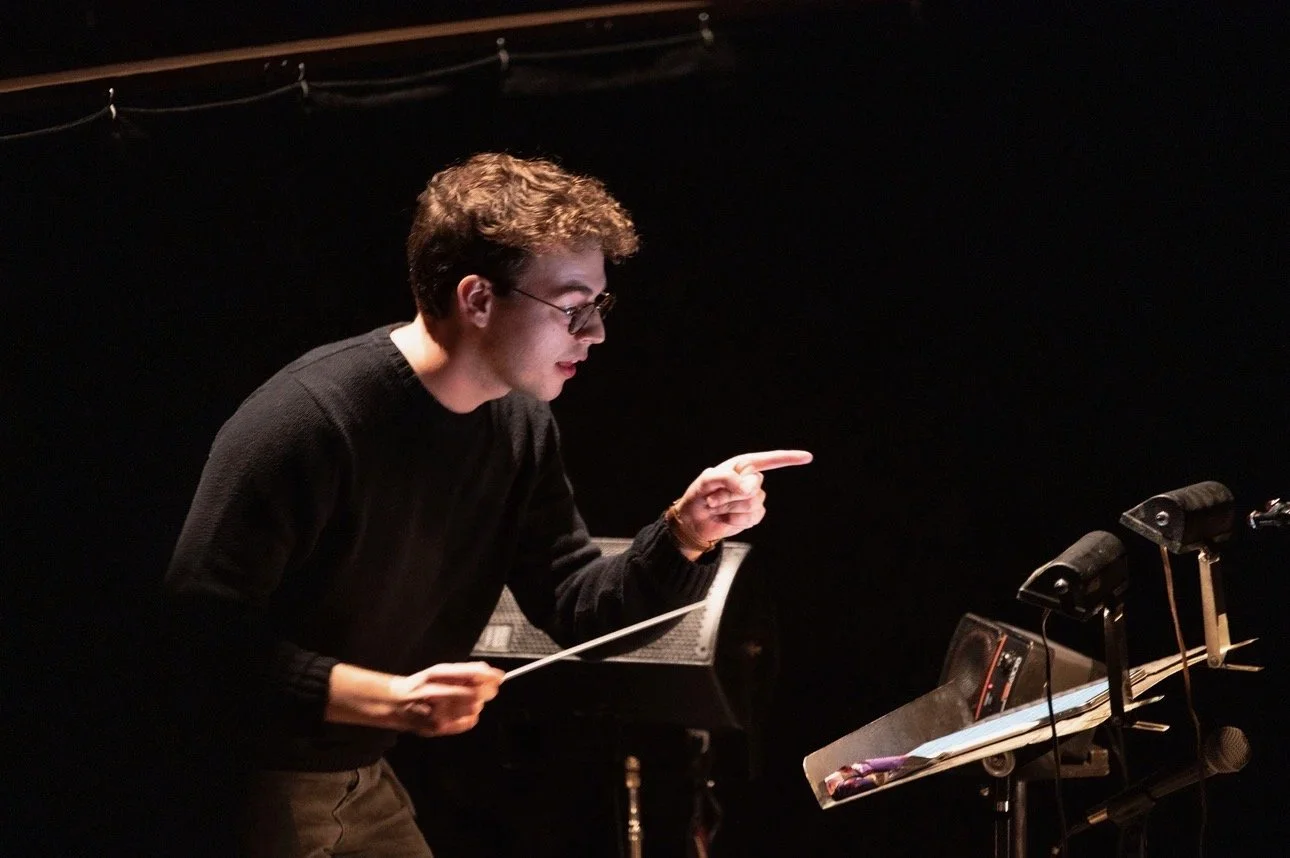Andrew conducting an orchestra or choir, gesturing with his right hand, holding a baton in his left hand, standing in front of music stands with illuminated sheet music, in a dark space with stage lighting.