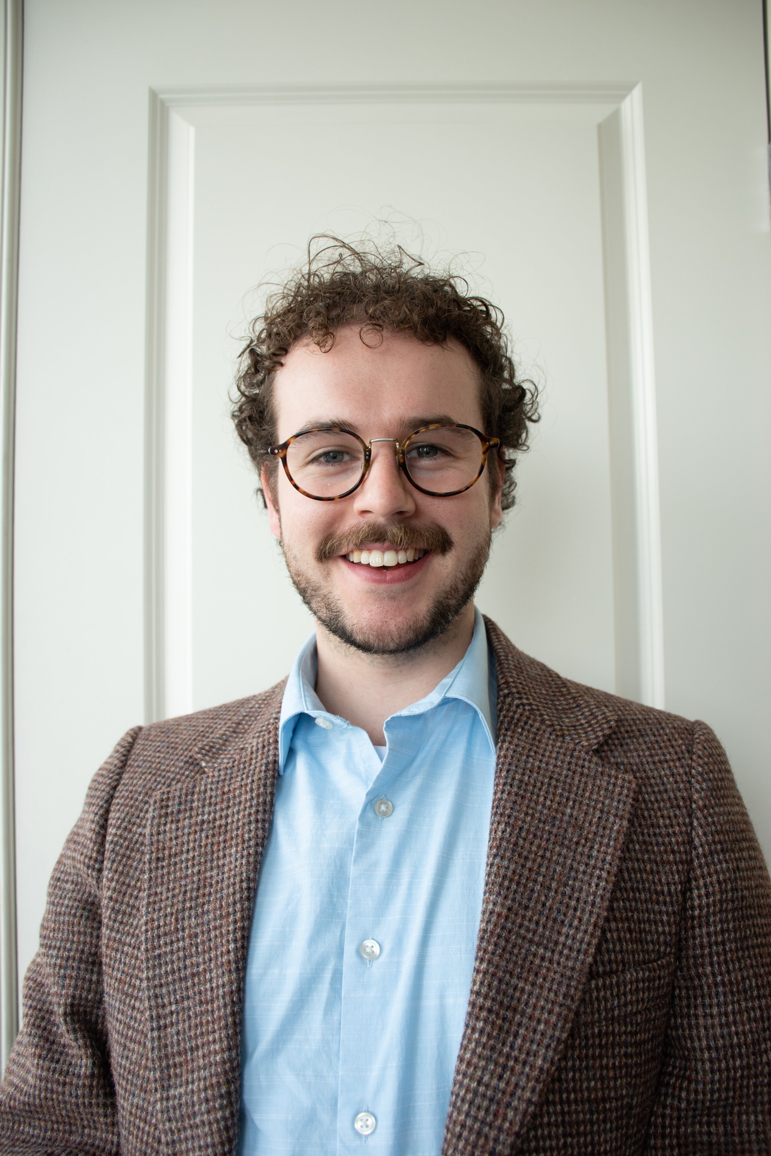 A young man (Andrew) with curly hair, glasses, and a beard, smiling, wearing a blue shirt and a brown plaid blazer, standing in front of a plain white door.