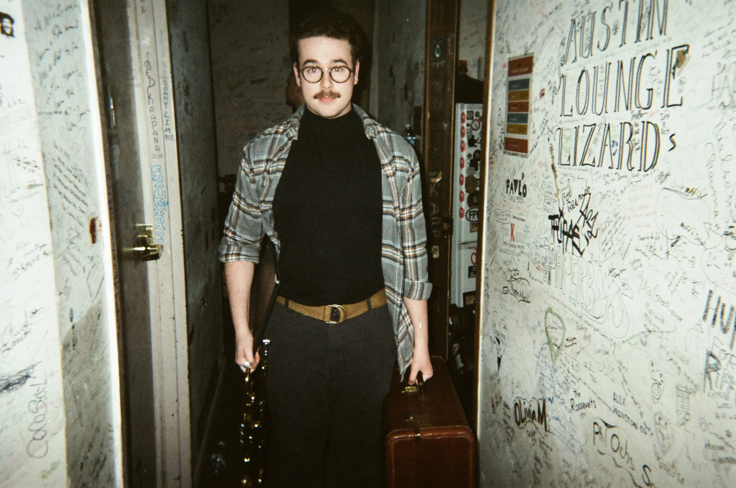 A man with curly hair, glasses, a black turtleneck, and a plaid shirt standing in a corridor covered with graffiti and signatures. He is holding a trumpet and a briefcase.