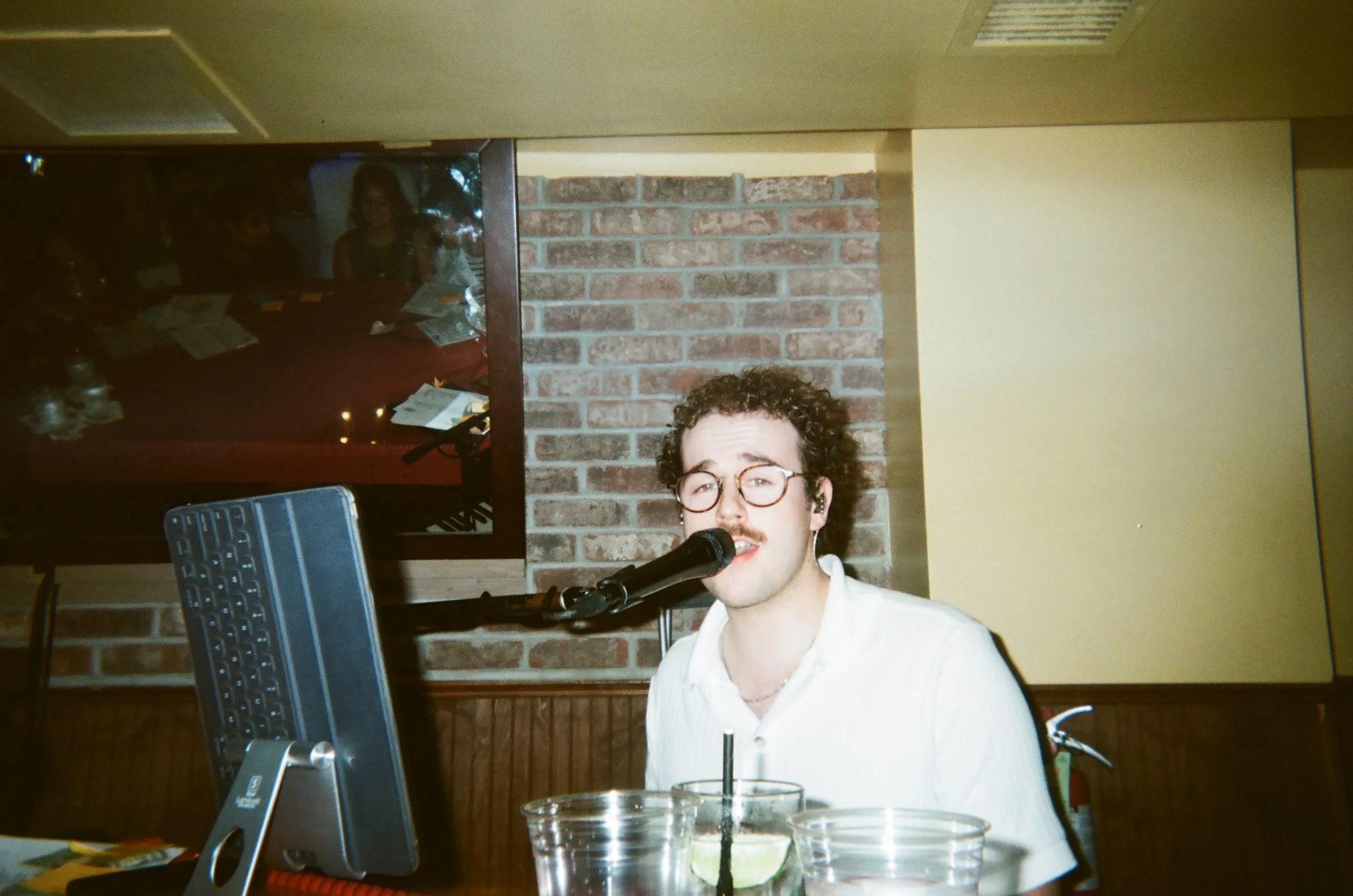 Andrew with curly hair, glasses, and a mustache singing into a microphone at a bar or restaurant. There are drinks with lime slices on the table and a laptop in front of him, with a brick wall and a glass window showing two women in the background.