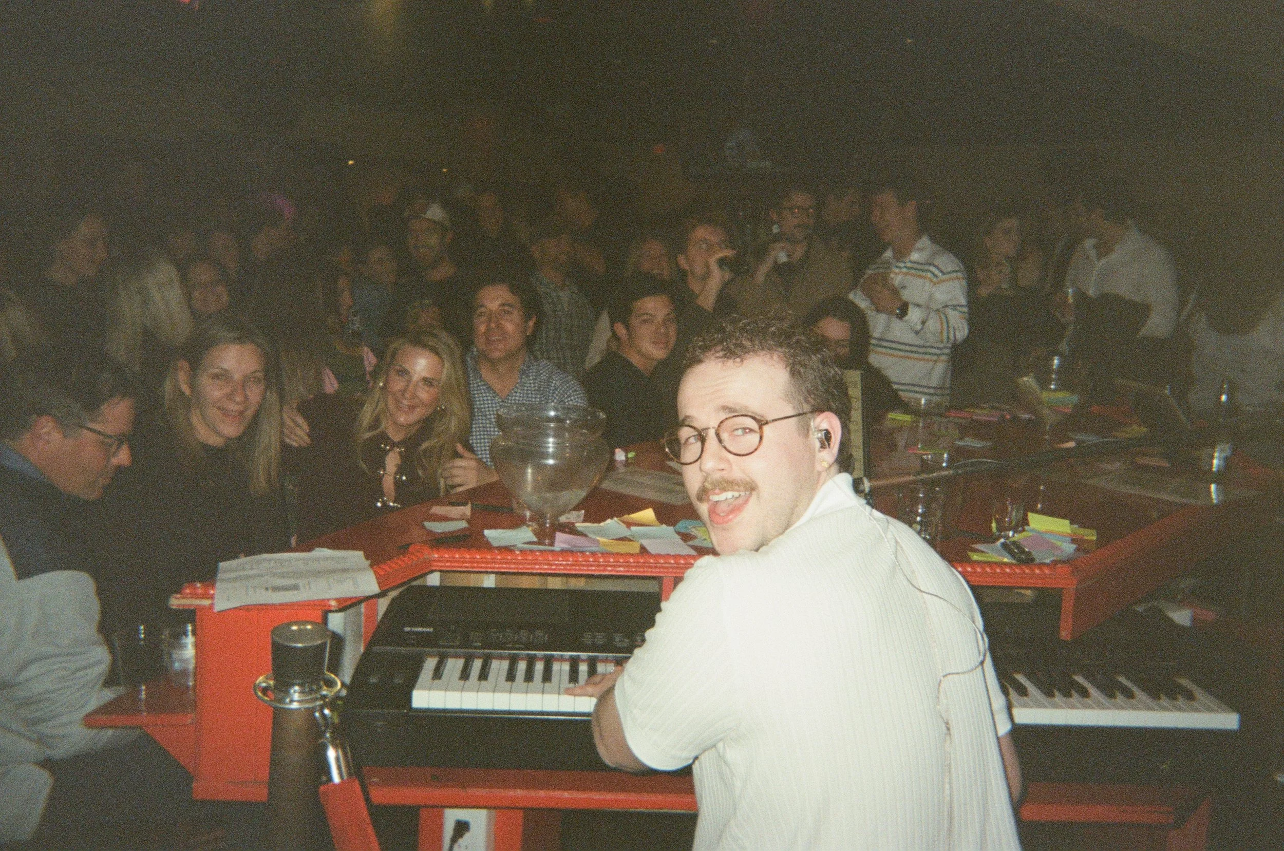 A group of people gathered at a party with Andrew playing a keyboard in the foreground.