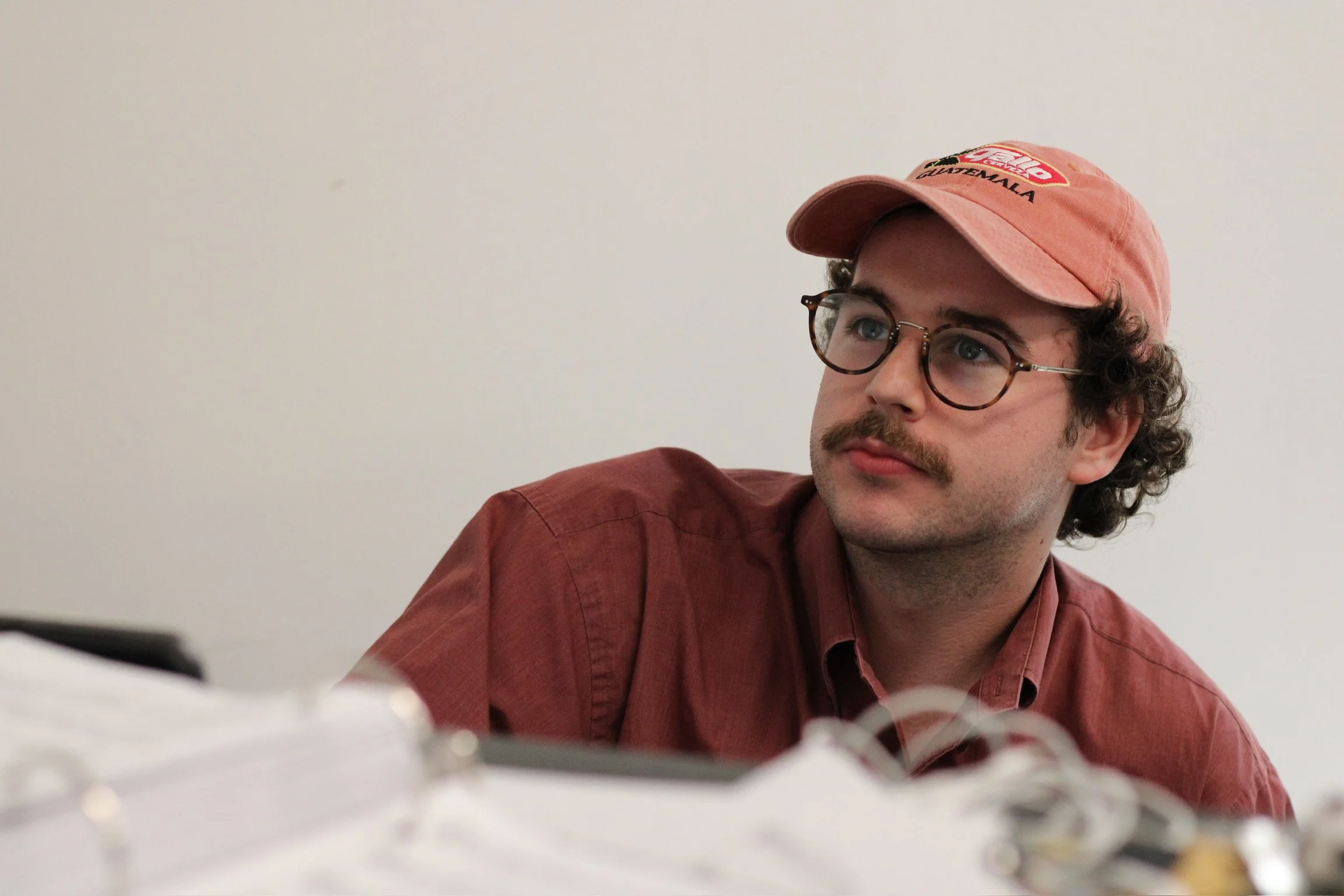 A young man with curly hair, glasses, and a mustache, wearing a pink cap and a maroon shirt, (Andrew) is seated against a plain white wall, looking to his left.