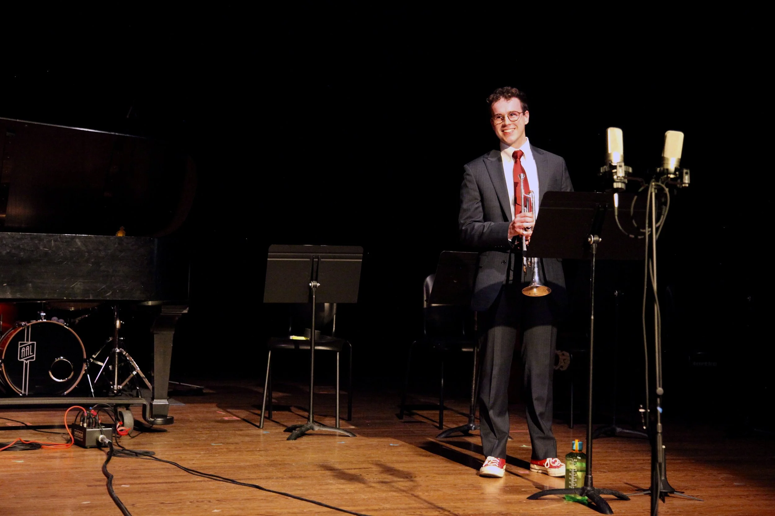 A young man in a suit and red tie holding a trumpet on stage with a smile, surrounded by musical instruments like a drum set and music stands.