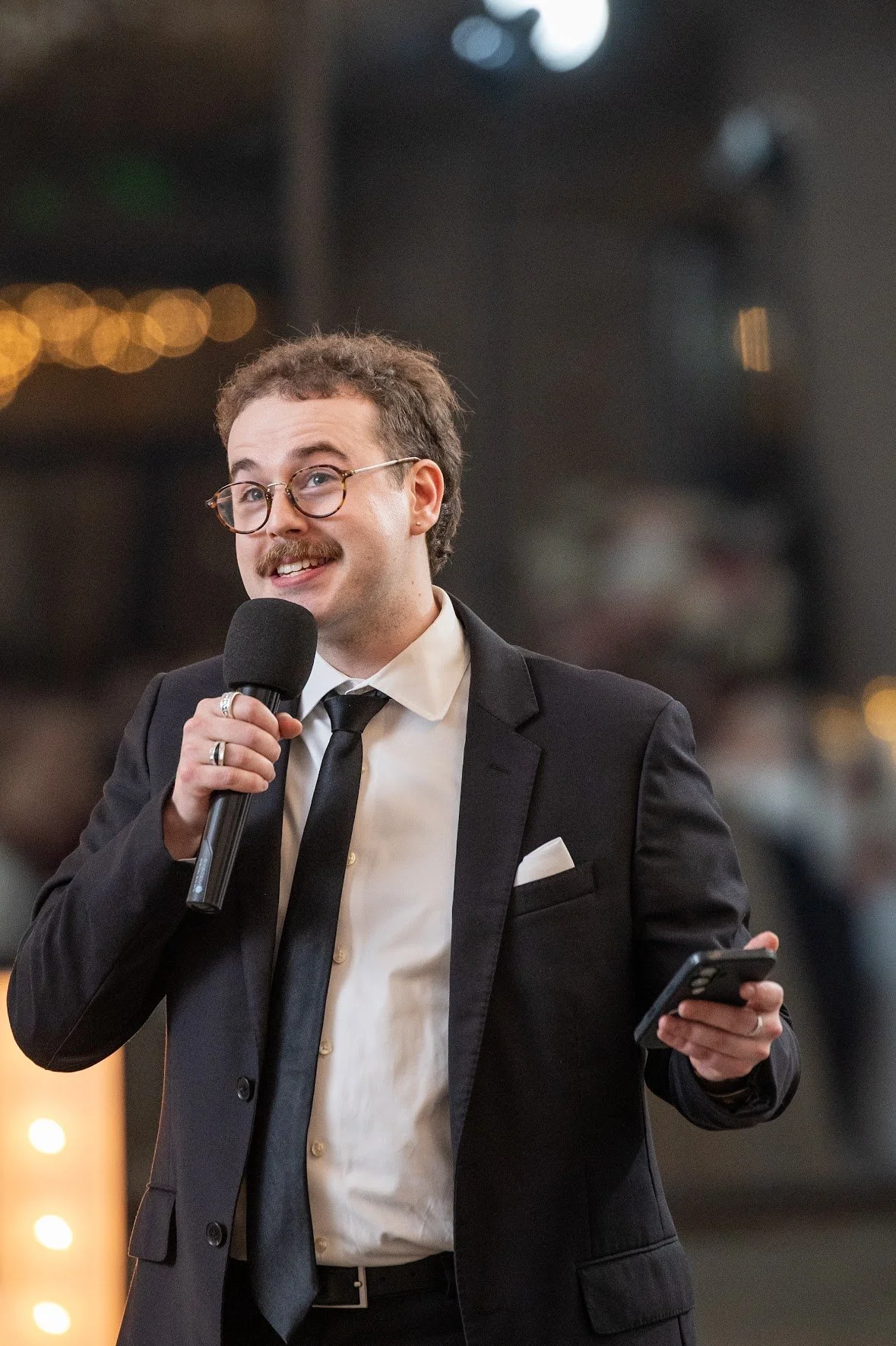 A man in a black suit, white shirt, and black tie, with glasses and a mustache, holding a microphone in one hand and a smartphone in the other, speaking at an event in a room with blurred lights and audience in the background.