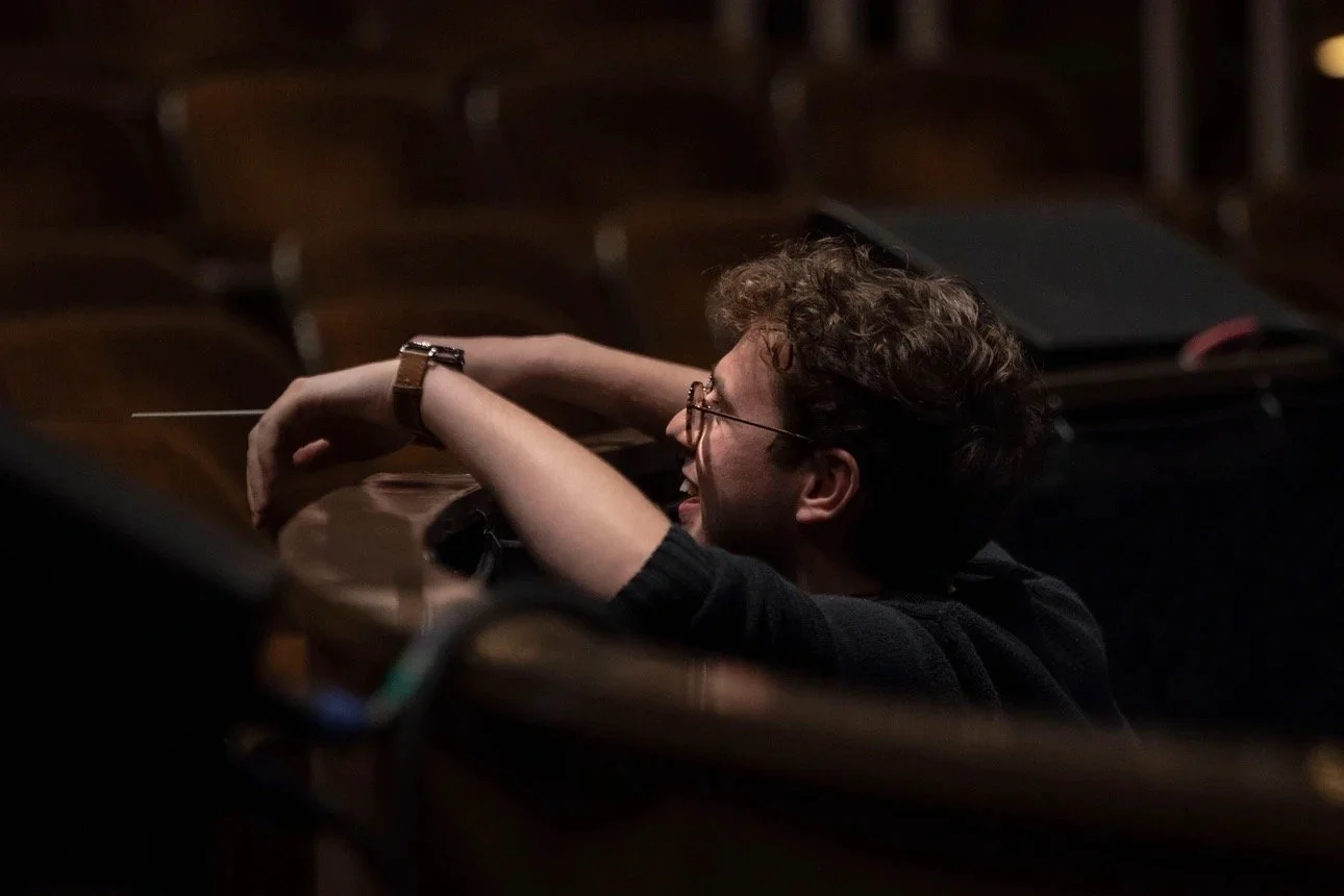 A person with curly hair and glasses (Andrew) resting in a dark auditorium, smiling with arms crossed on the ledge of the pit.