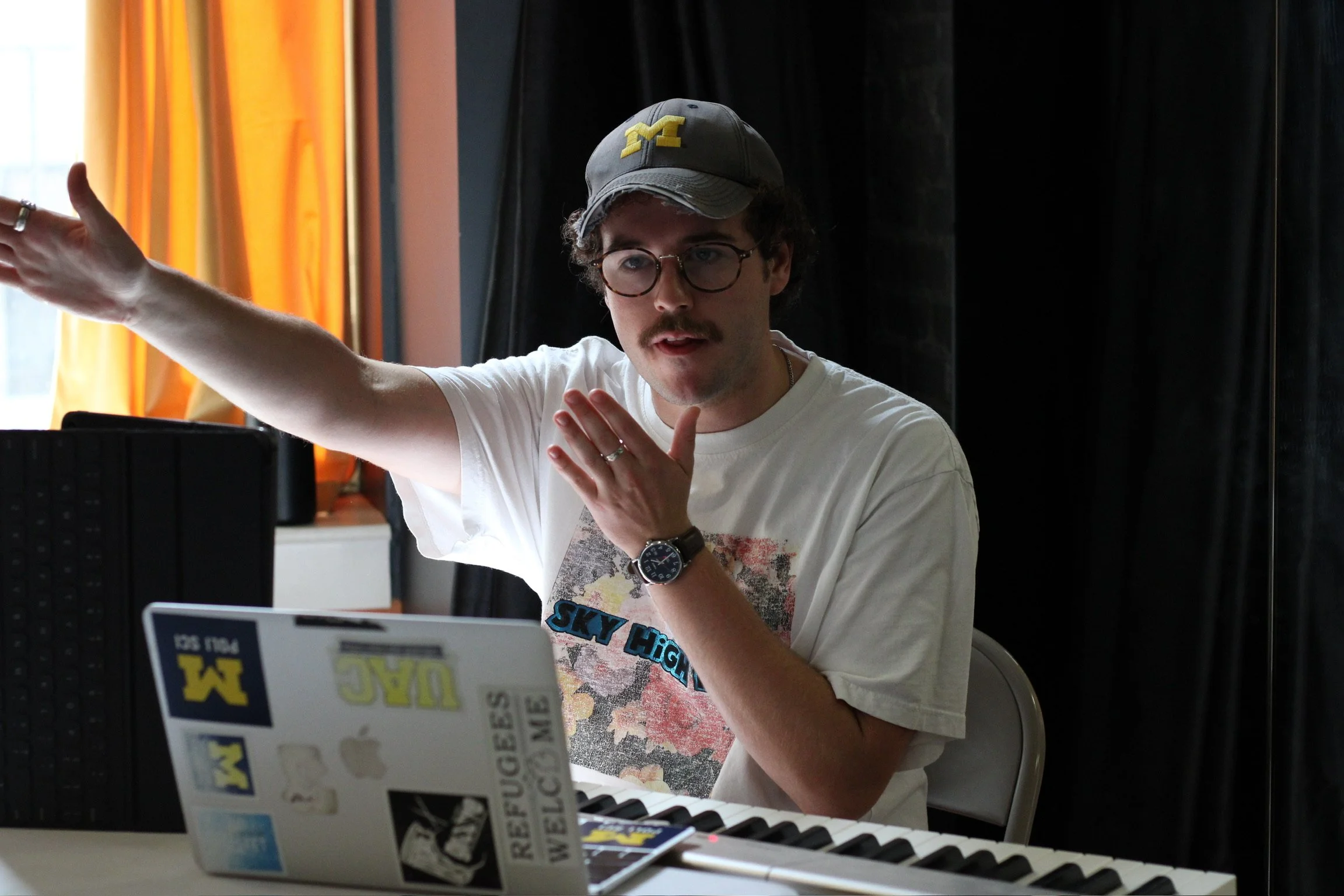 A young man with glasses and a mustache wearing a baseball cap and a graphic t-shirt sitting at a desk with a keyboard, wall, curtains, and a window in the background, gesturing with his left hand.