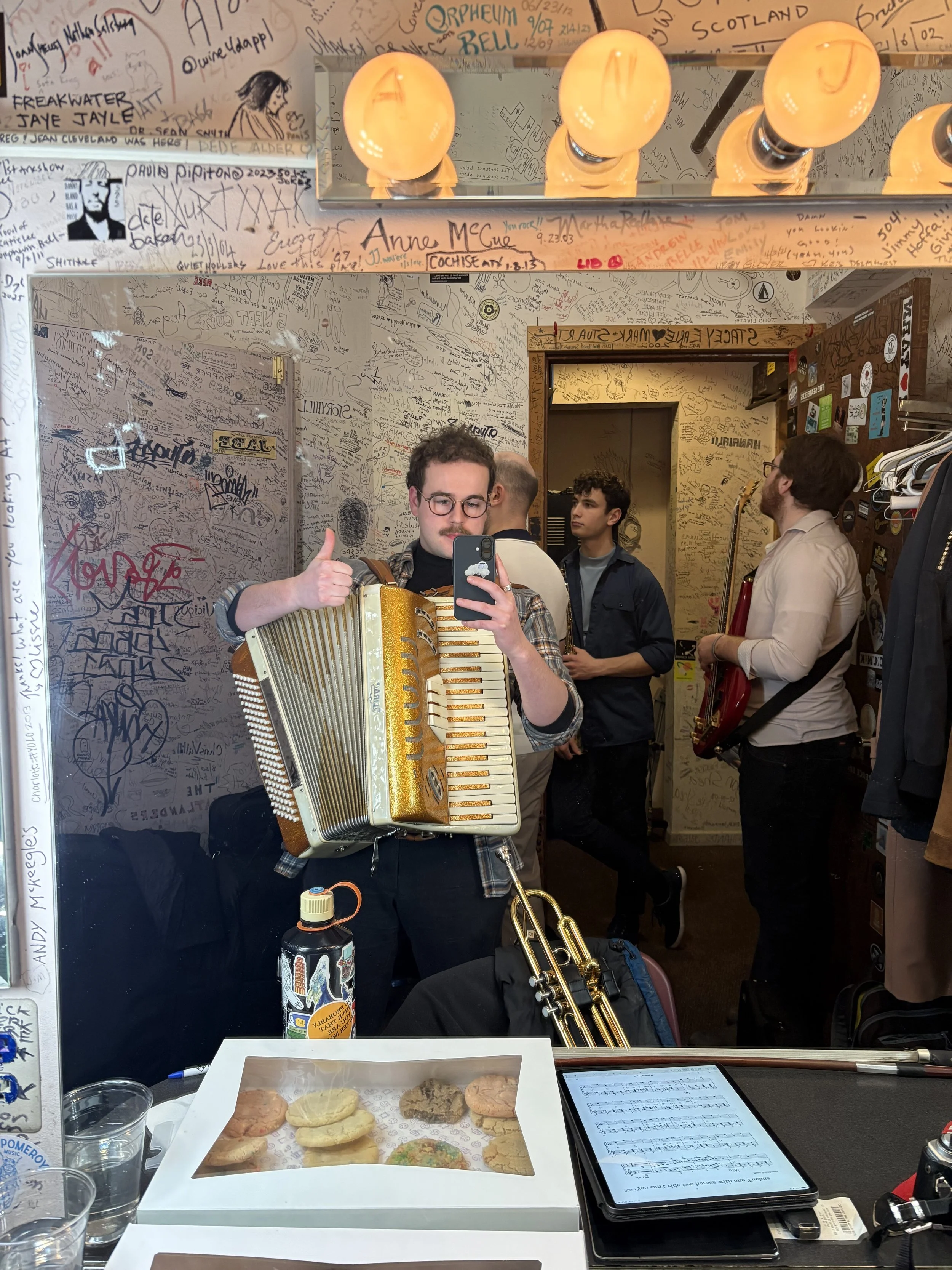 A person holding an accordion and giving a thumbs-up mirror selfie in a room with graffiti-covered walls and ceiling, with other people in the background. There are cookies in a white box, a water bottle, glasses of water, a music sheet on a tablet, 