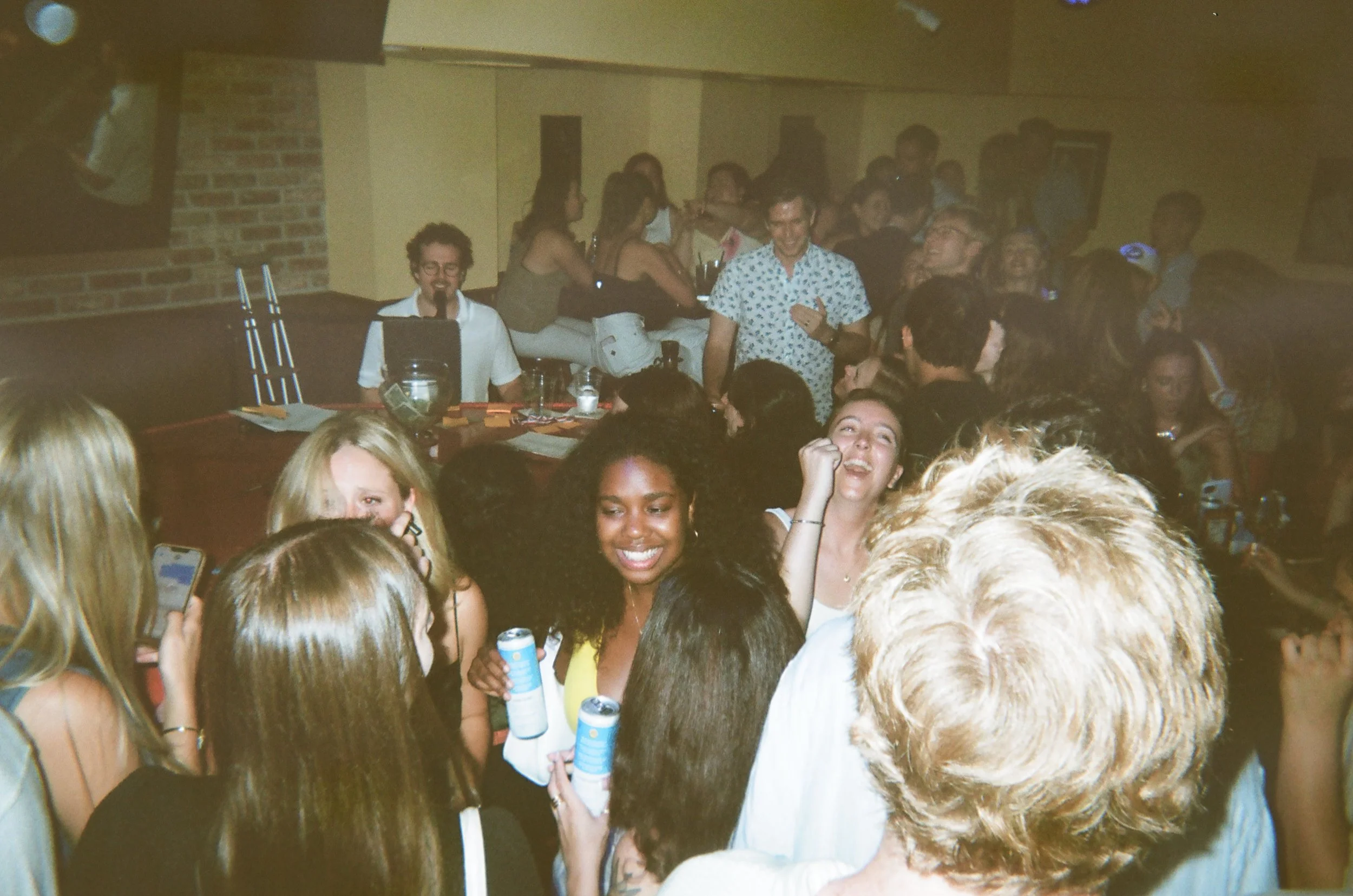 Group of people at a party or gathering, some smiling and holding drinks, with a man in a floral shirt smiling and a woman in a yellow top in the foreground.