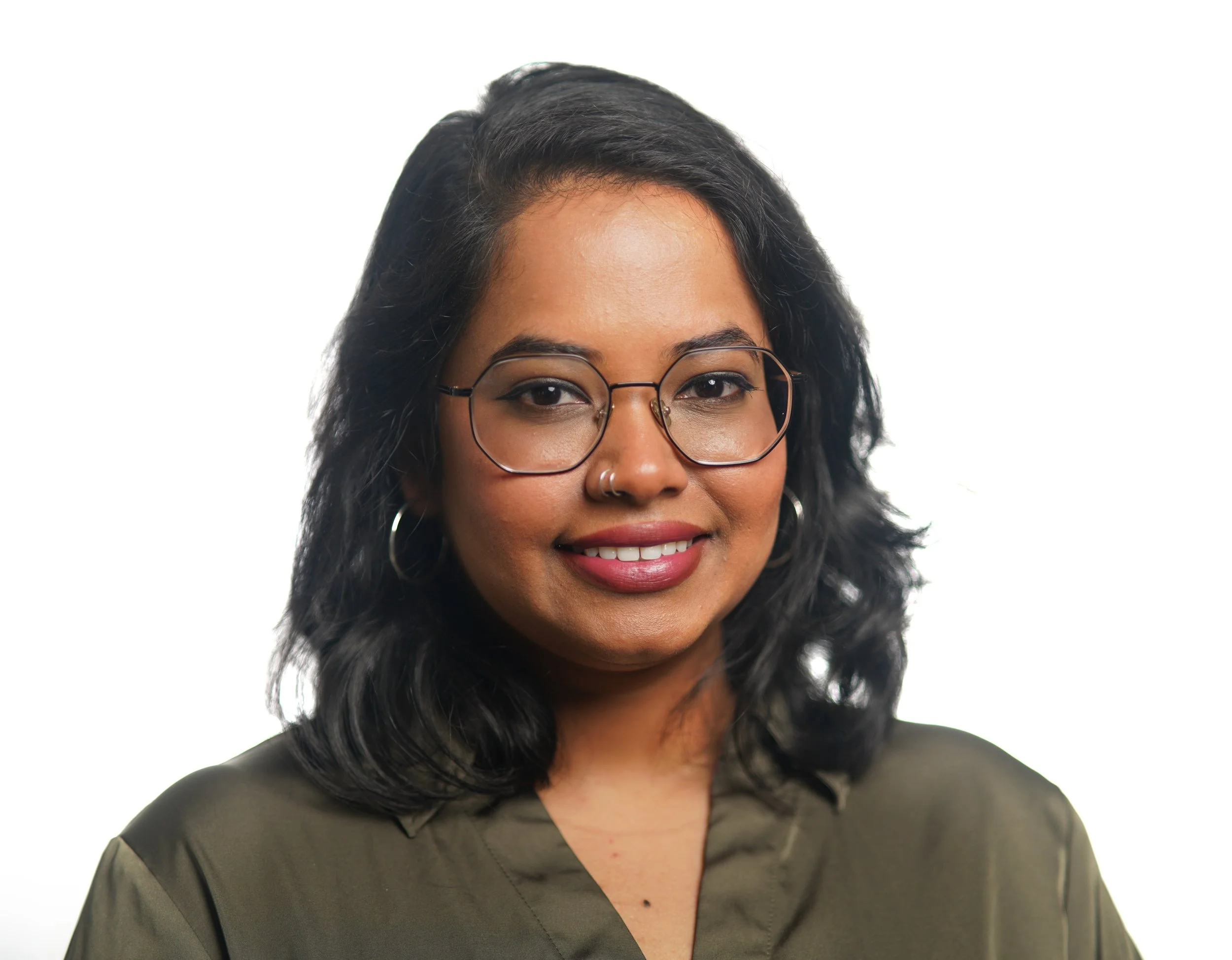 Close-up of a woman with medium skin tone, shoulder-length curly black hair, wearing glasses, earrings, and a black top, smiling against a plain white background.