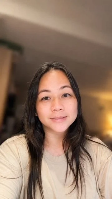 Young woman with long dark hair and a beige top taking a selfie in a home setting.