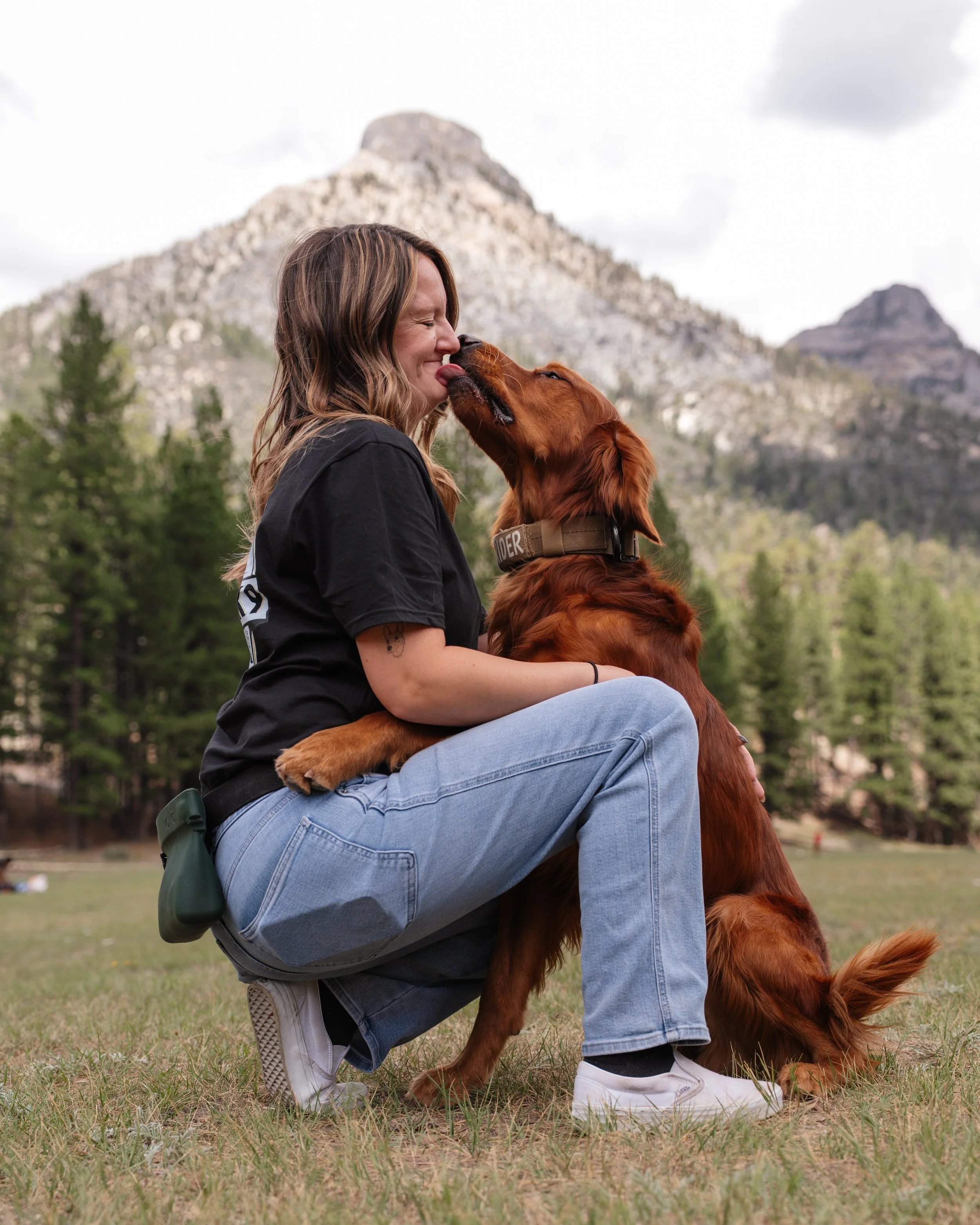 A woman with wavy hair wearing a black T-shirt and light blue jeans squatting on grassy ground, playing with her golden retriever puppy outdoors in a park with tall trees and a mountain in the background.