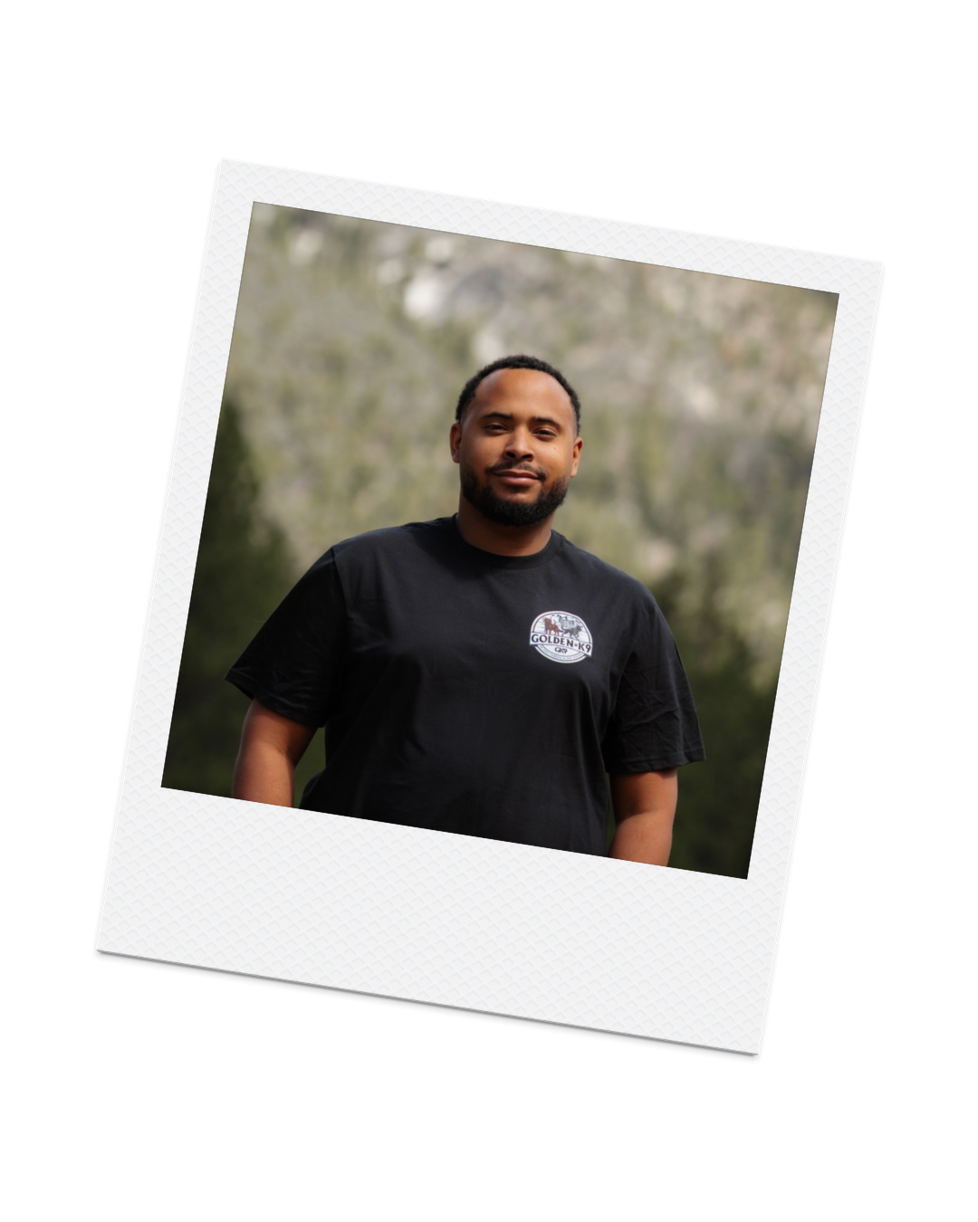 Portrait of a man with a beard and short hair, wearing a black T-shirt with a logo, standing outdoors with blurred trees in the background.