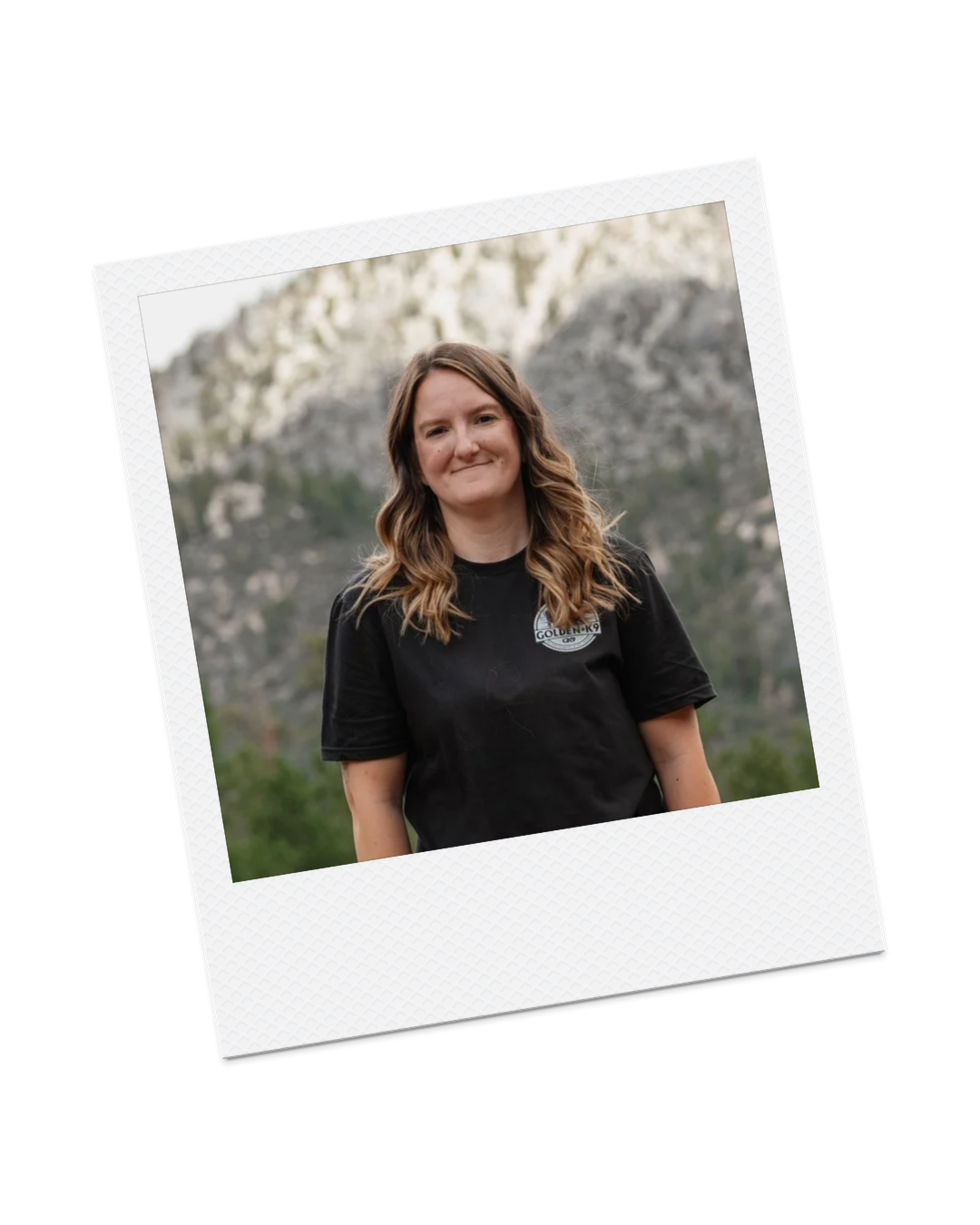 A woman with wavy, shoulder-length brown hair smiling outdoors with mountainous scenery in the background, wearing a black T-shirt with a patch on the chest.