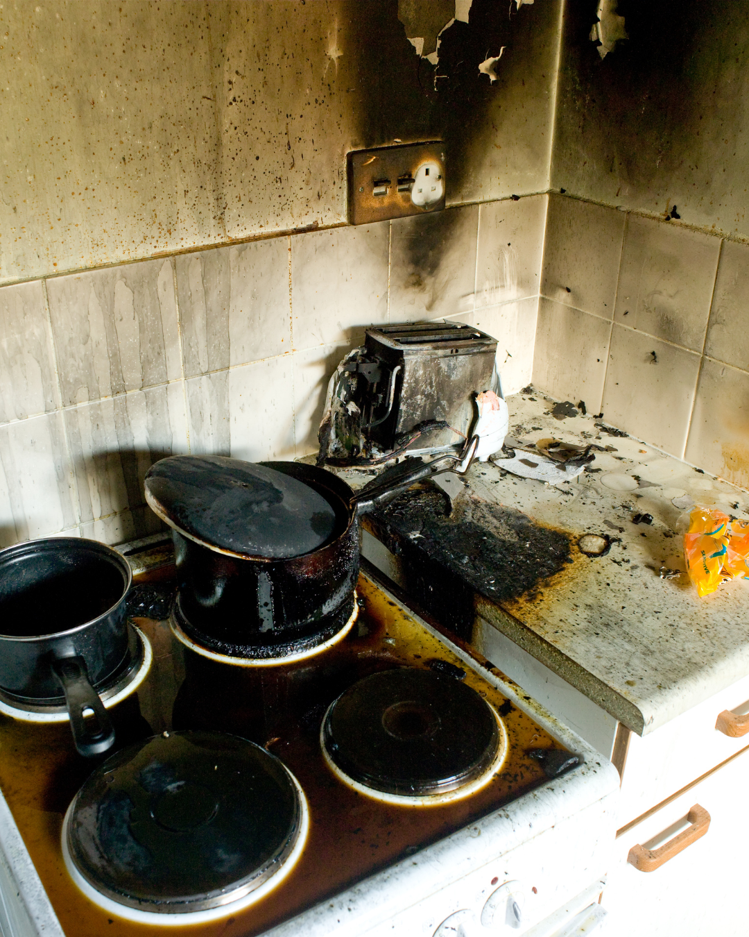 Kitchen stovetop and wall with severe fire damage; burnt pots, charred stove surface, blackened wall and backsplash.