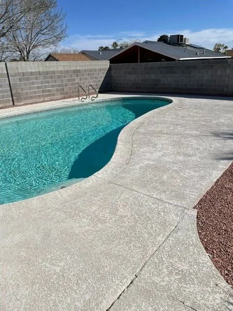 Residential backyard pool with concrete deck, surrounded by a brick wall and a house in the background under a blue sky.