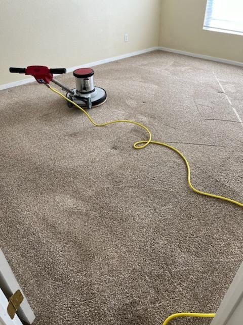 A carpet cleaning machine on a beige carpet in a room with beige walls and a window with blinds.