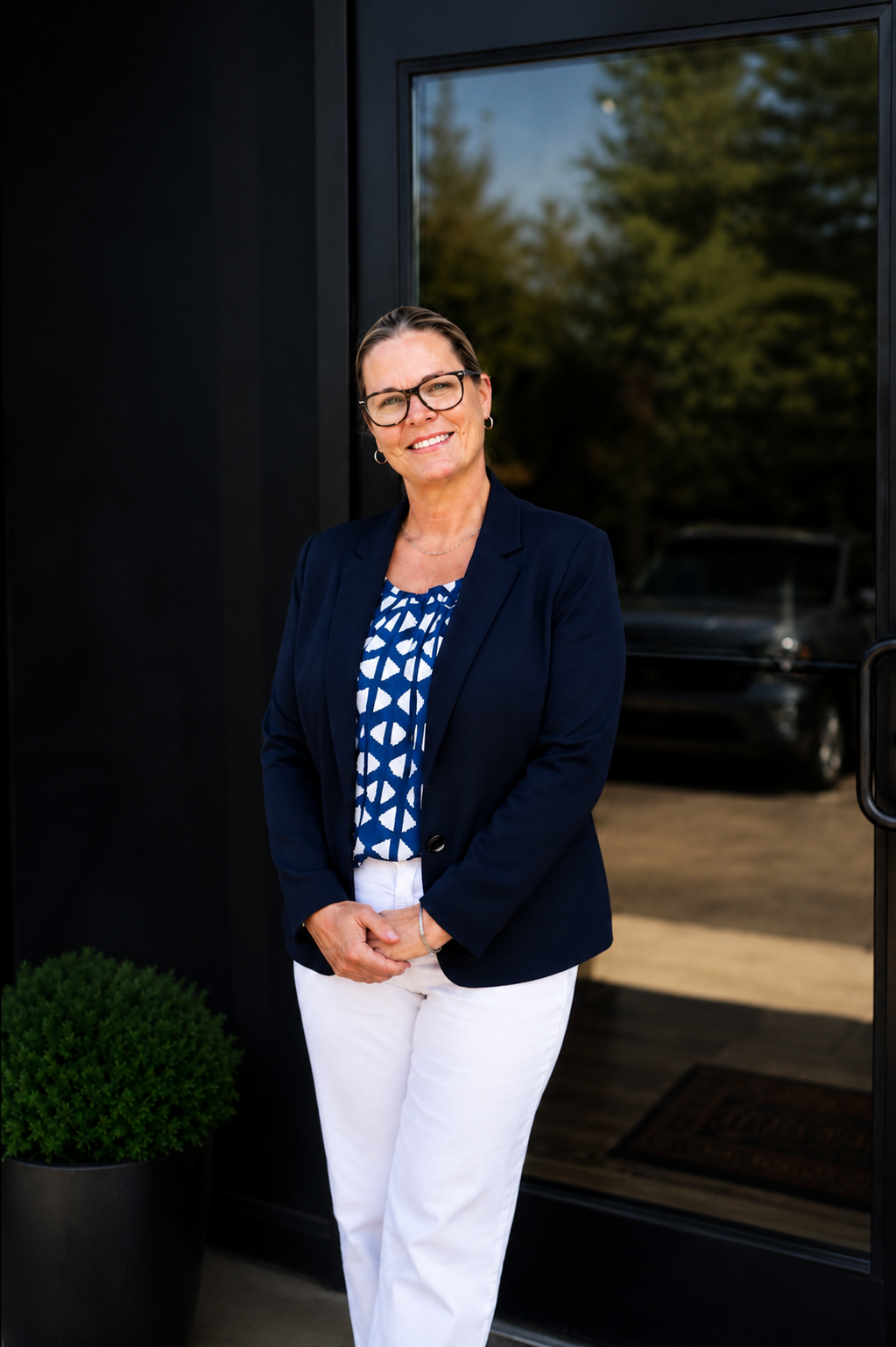 A woman with glasses in a navy blazer, patterned blouse, and white pants stands outside a modern glass door, smiling at the camera.