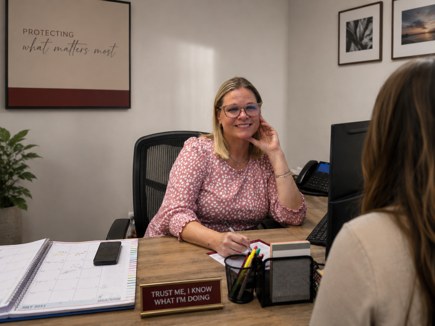 A woman with blonde hair, glasses, and a pink polka-dot blouse is sitting at a desk, smiling at the camera. She is holding a pen, with a calendar, phone, and office supplies on her desk. There is a sign that reads, "Trust me, I know what I'm doing." Behind her, there are framed wall art and a potted plant.