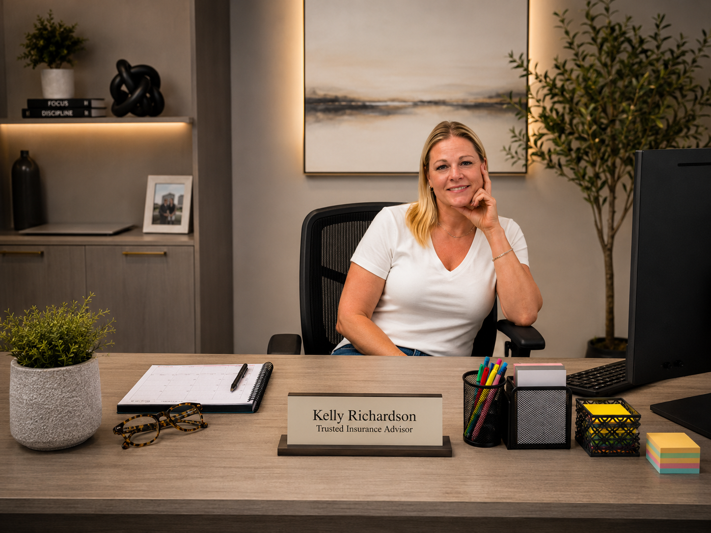 A woman smiling and sitting at a desk in an office environment. She has blonde hair and is wearing a white shirt. There is a nameplate on the desk that reads "Kelly Richardson, Trusted Insurance Advisor." The desk has various items including a potted plant, glasses, a notebook with a pen, colorful markers, sticky notes, and a computer monitor. There are shelves behind her with decorations, books, and a framed photo, along with a painting on the wall and a large plant in the corner.