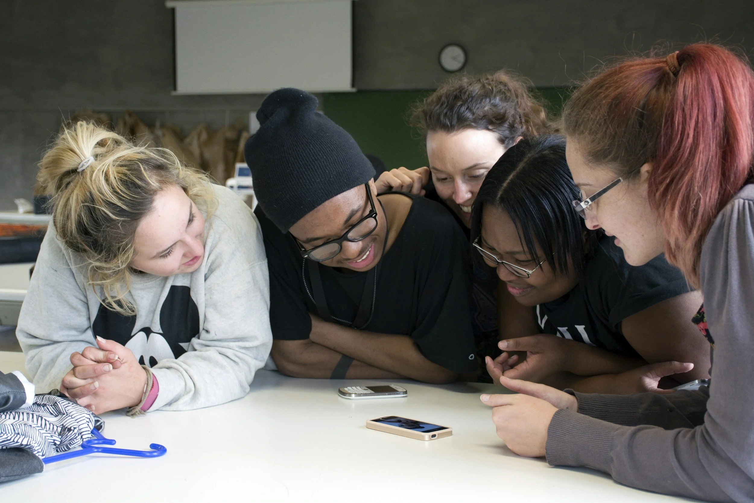 Group of five young women gather around a phone on a table, looking at it and smiling, in a classroom setting.