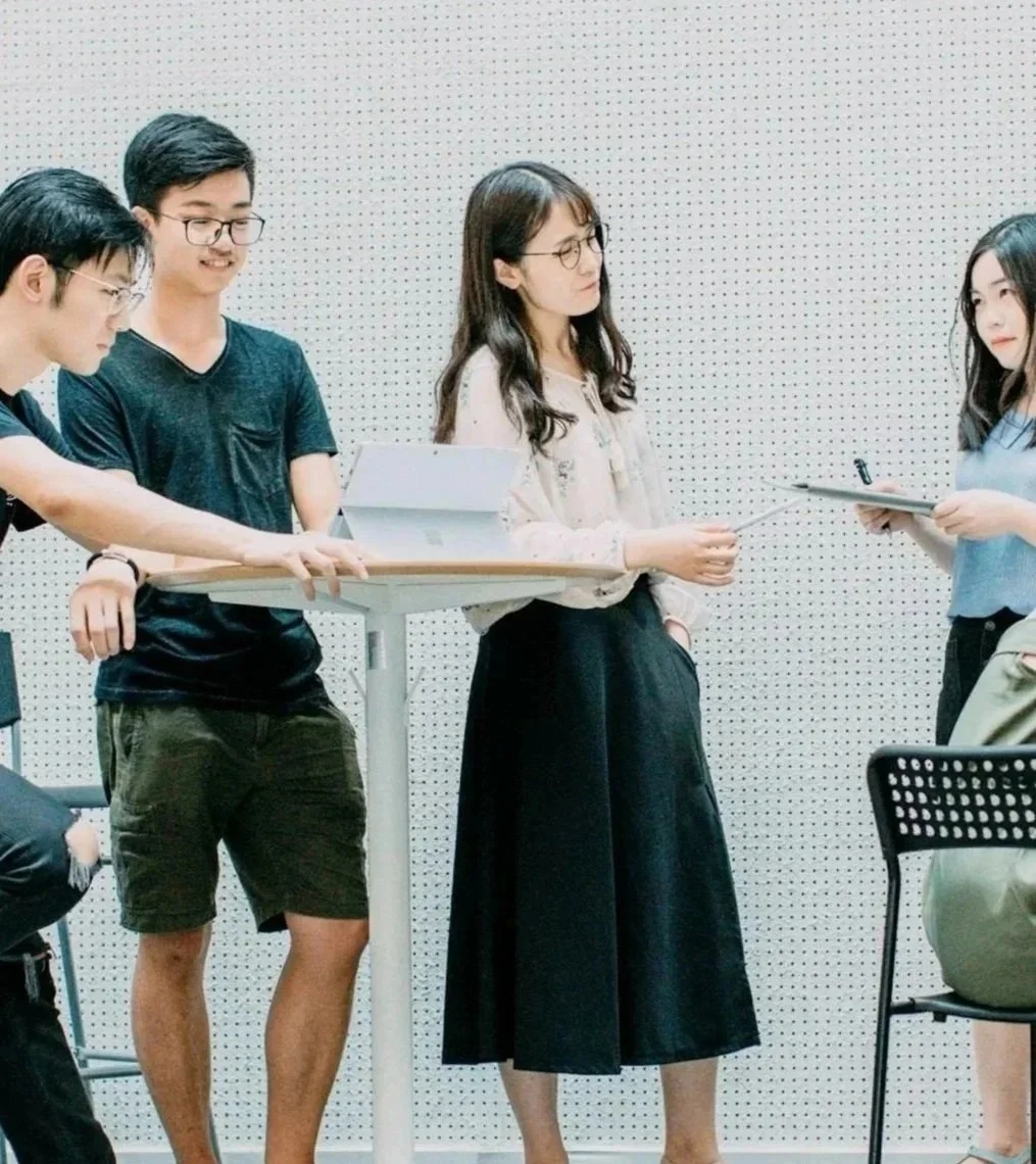 Group of four young Asian individuals in discussion; one woman holding a document, others listening, in a room with white perforated walls.