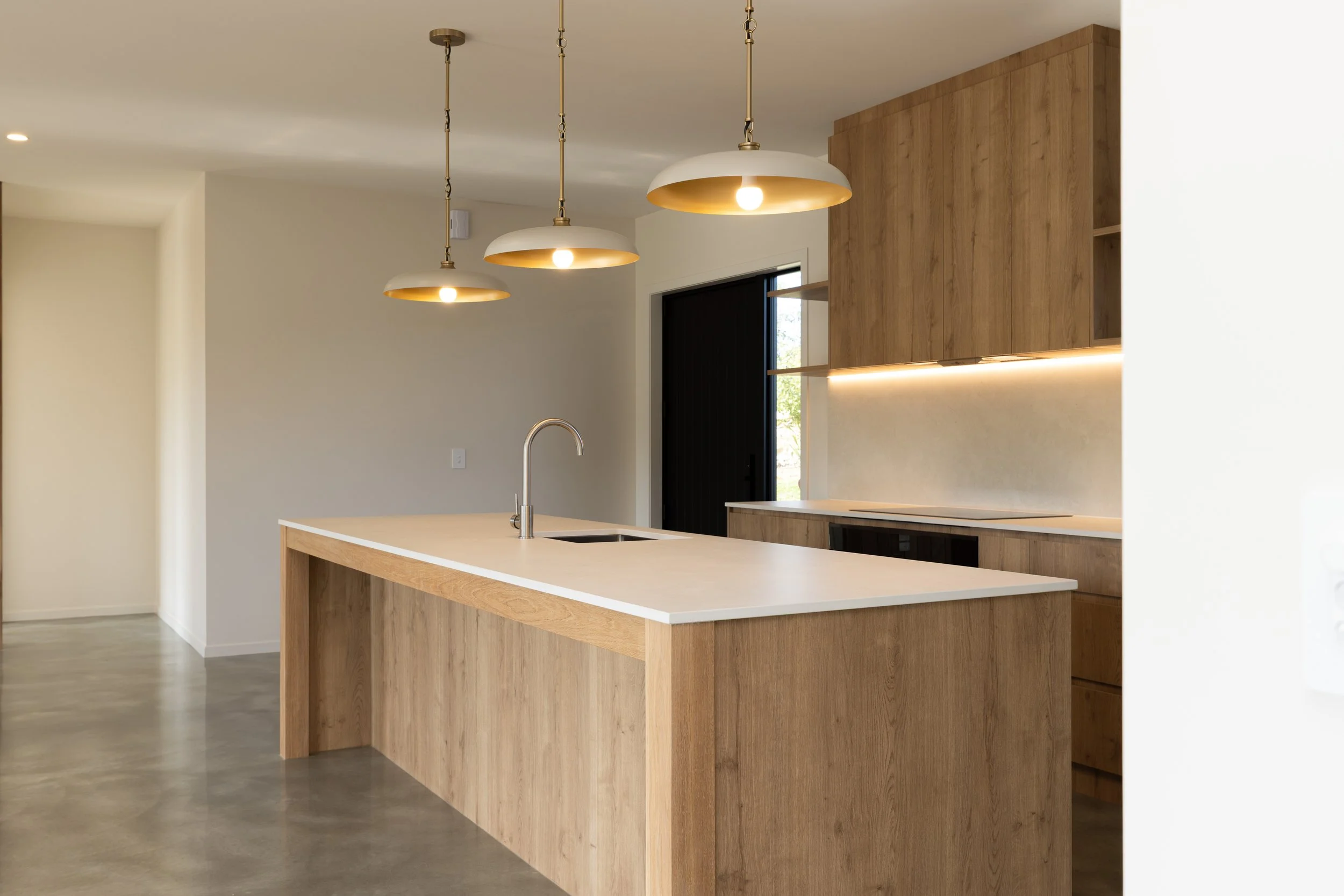 Modern kitchen with a white countertop island, wooden cabinets, and three pendant lights hanging from the ceiling, near a black door and a window.