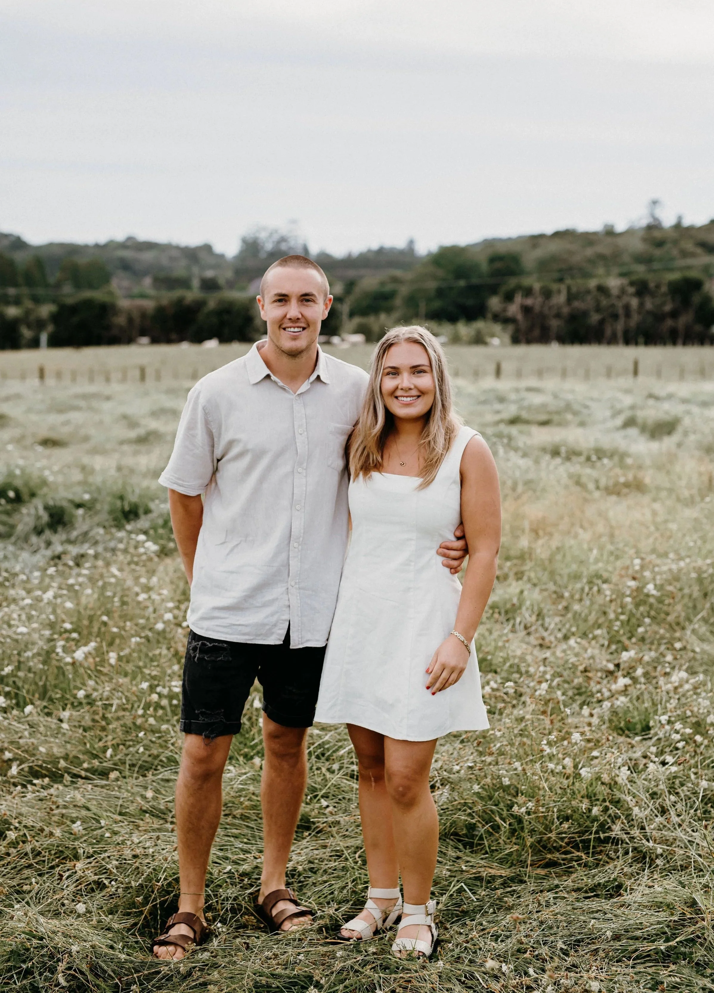 A young man and woman stands together outdoors in a field, smiling at the camera. The man is wearing a light-colored button-up shirt, black shorts, and brown sandals. The woman is wearing a white dress, white high-heeled sandals, and a bracelet on her left wrist. The background includes rolling hills and trees under a cloudy sky.