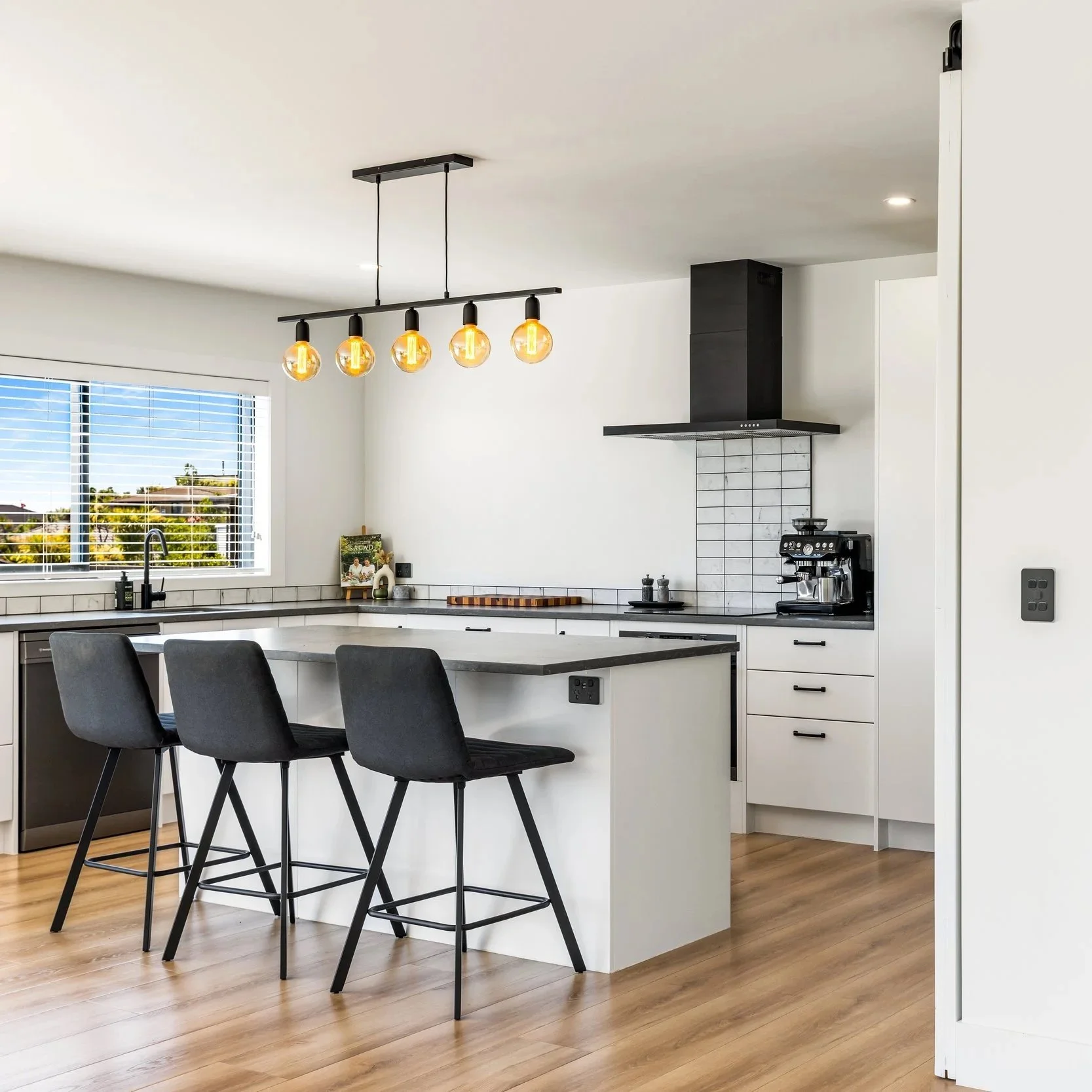 Modern kitchen with white cabinets, black countertops, a black range hood, and a black coffee machine on the counter. A large window with white blinds overlooks greenery outside. A black pendant light with five exposed bulbs hangs above a kitchen island with three black chairs. Light wood flooring completes the space.