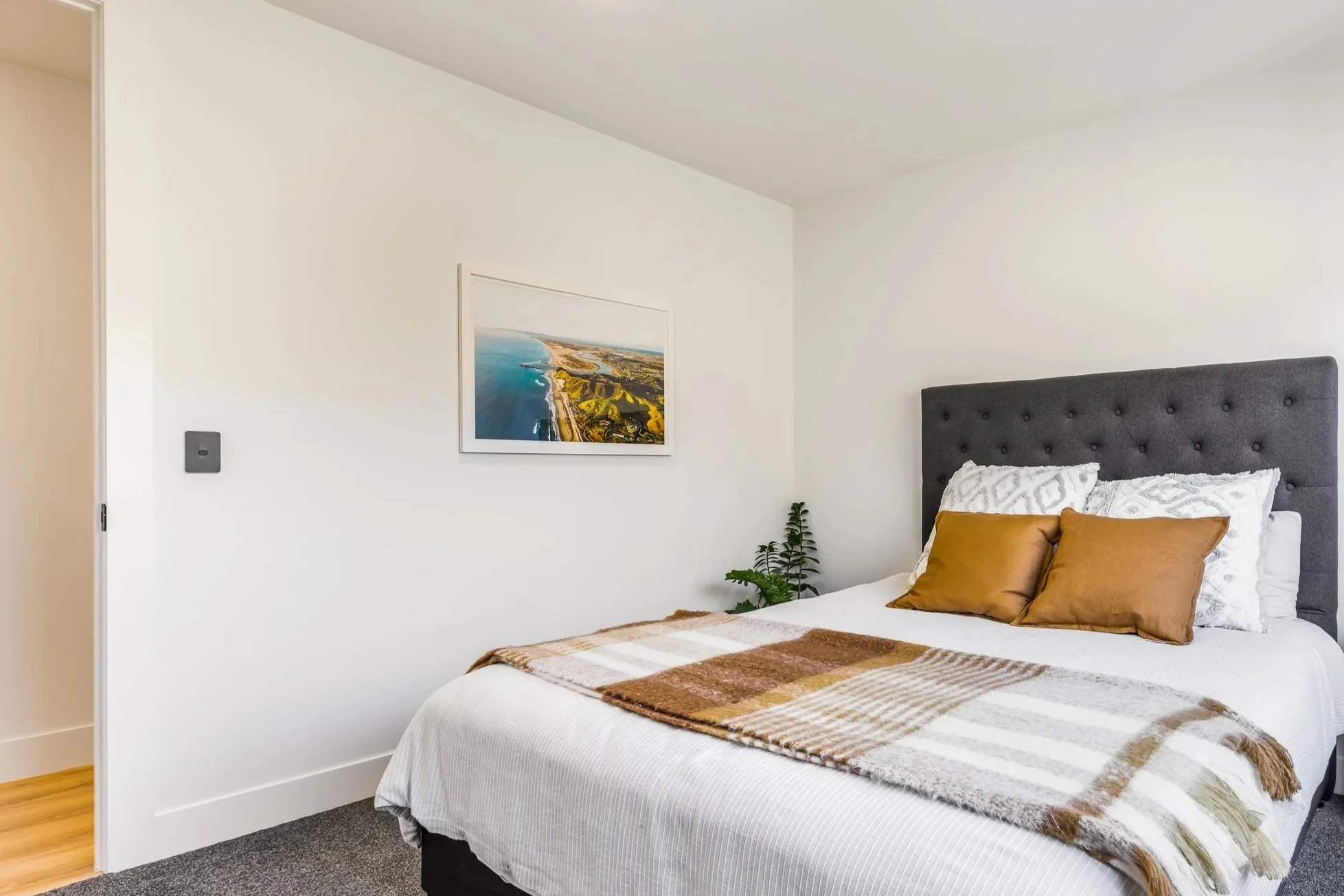 A bedroom with a bed having a gray headboard, white bedding, and brown pillows. There is a wall-mounted picture of a coastline, a small plant, and a light switch on the wall.