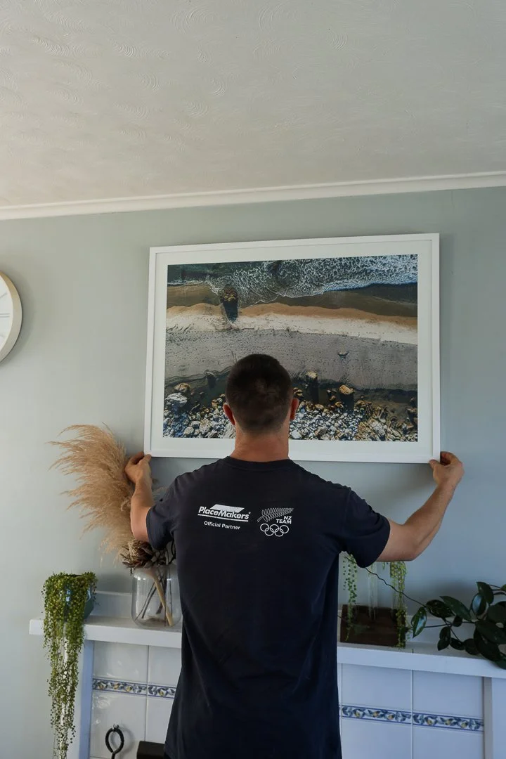 A man hanging a framed aerial beach photograph on a light gray wall in a room decorated with plants and a clock.