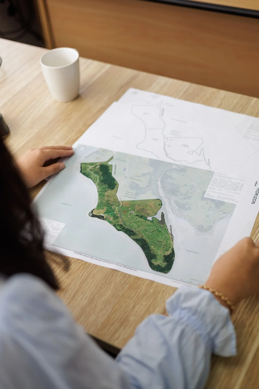 Person holding a map of a land development project, sitting at a wooden table with a blank white paper and a white cup nearby.