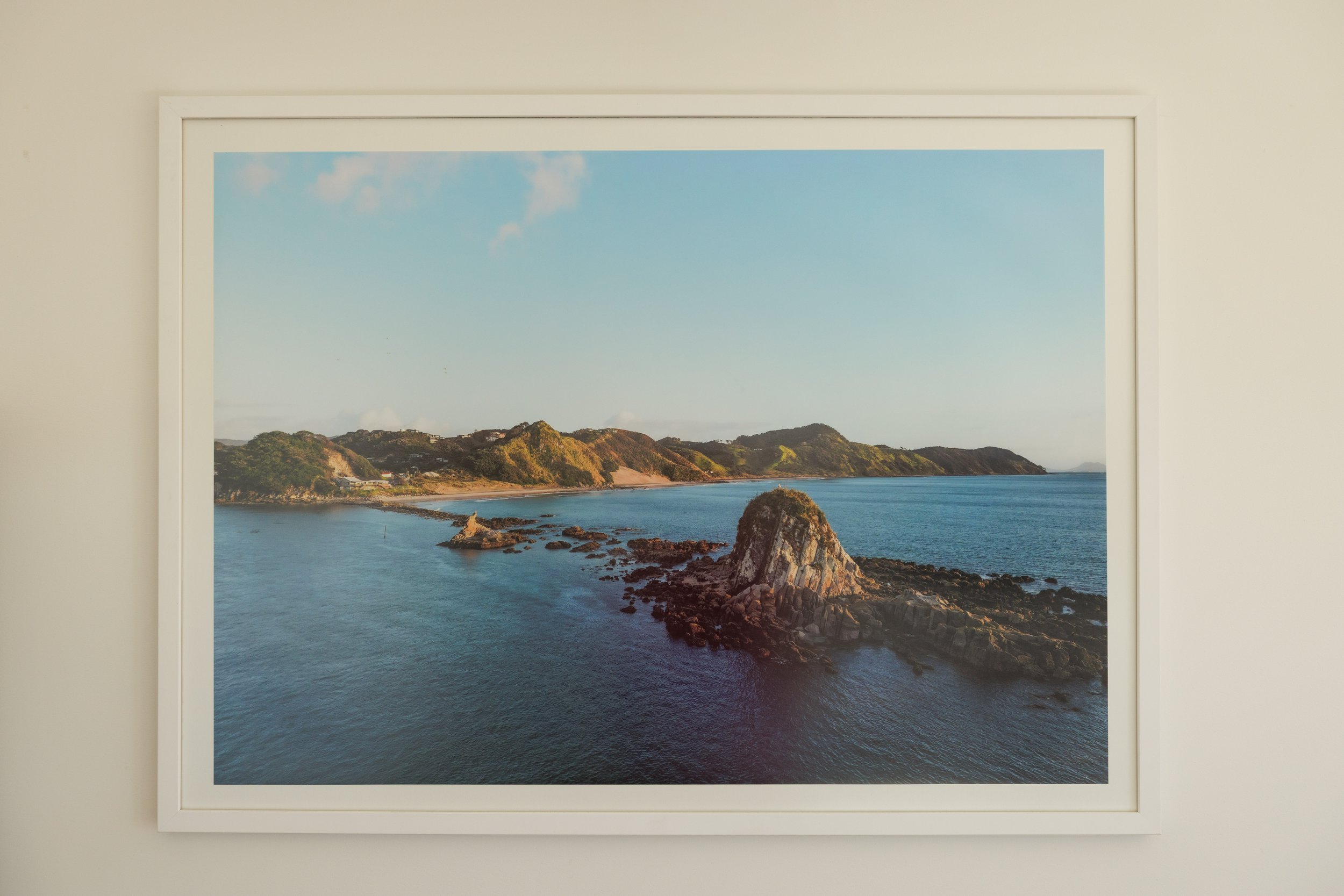 Framed landscape painting or photograph of a coastal scene with clear blue sky, green hills, rocky coastline, and calm water.