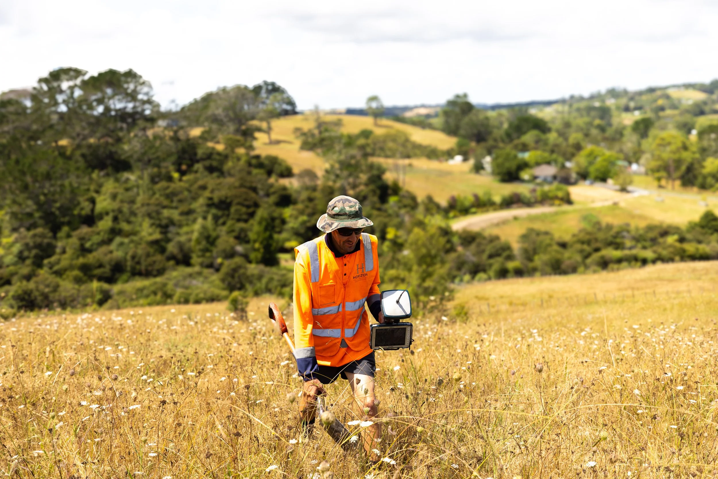 A man wearing a hat, sunglasses, and an orange safety vest is working in a field with tall grass and wildflowers. He is using a metal detector and holding a shovel. There is a landscape with rolling hills, trees, and houses in the background.