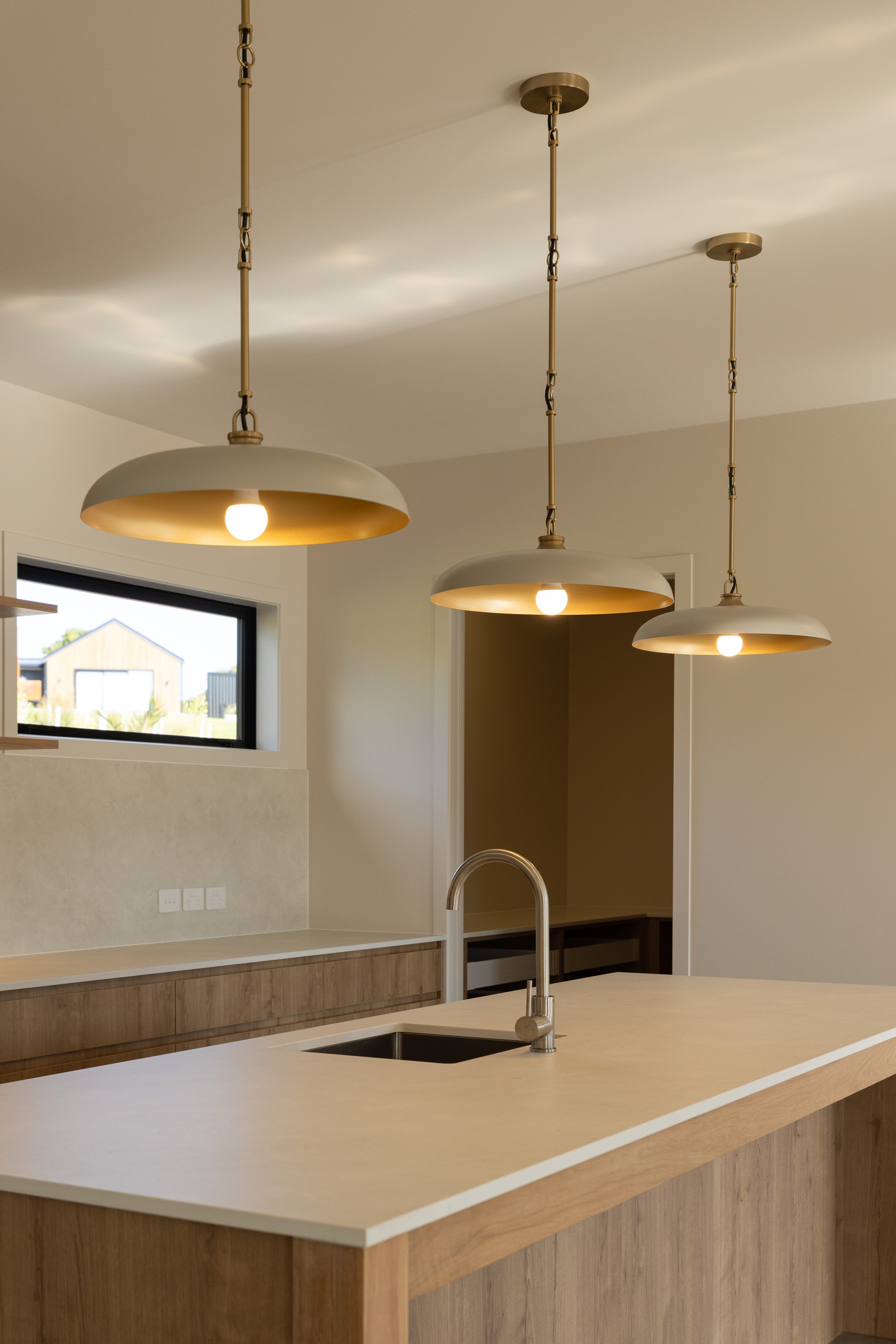 Modern kitchen island with a beige countertop, wooden base, and a stainless steel sink. Three pendant lights with white shades hang above the island. A small window in the background shows a neighboring building.