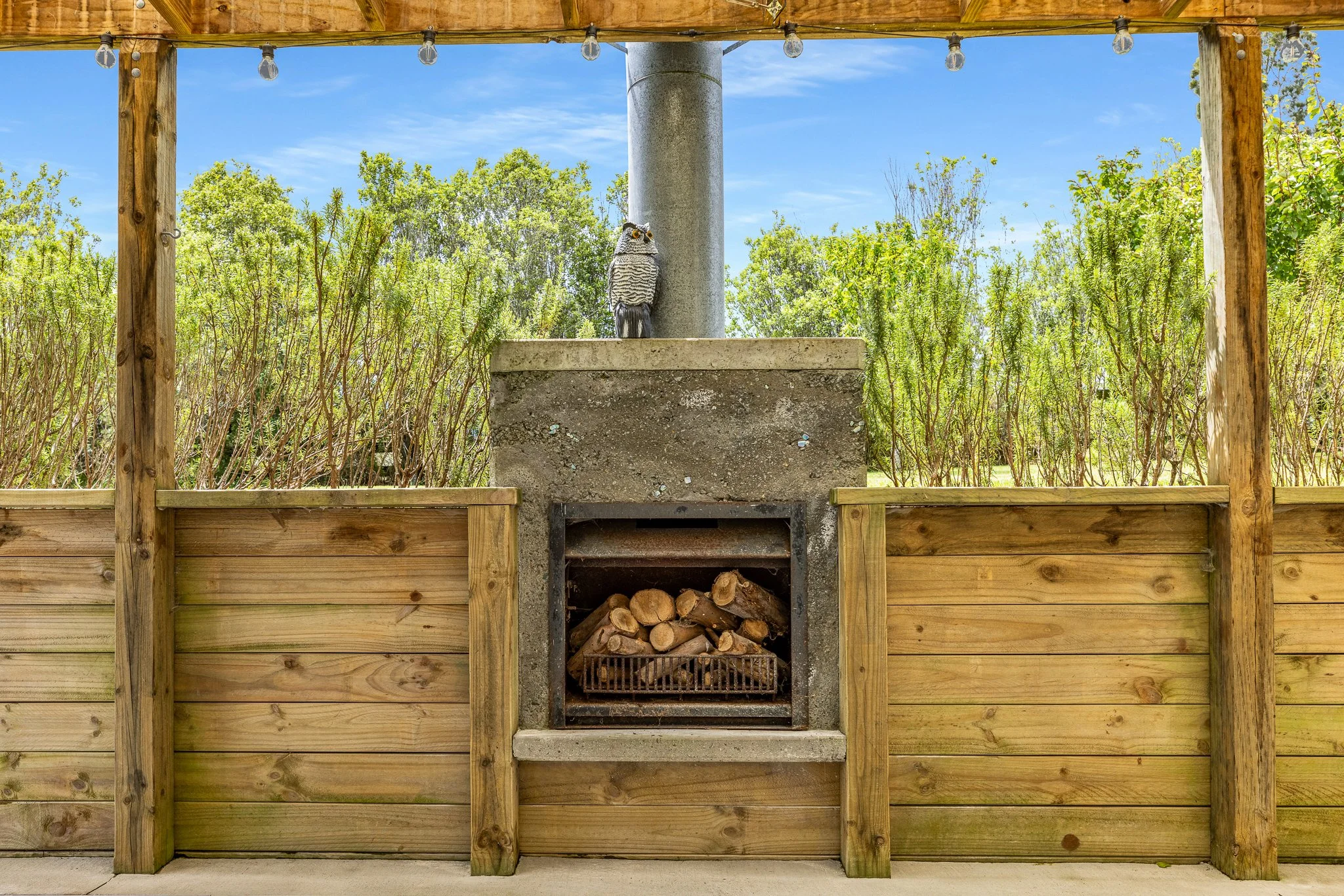 Wooden outdoor structure with string lights, a concrete fireplace with a woodpile underneath, and a decorative owl perched on a concrete pillar.