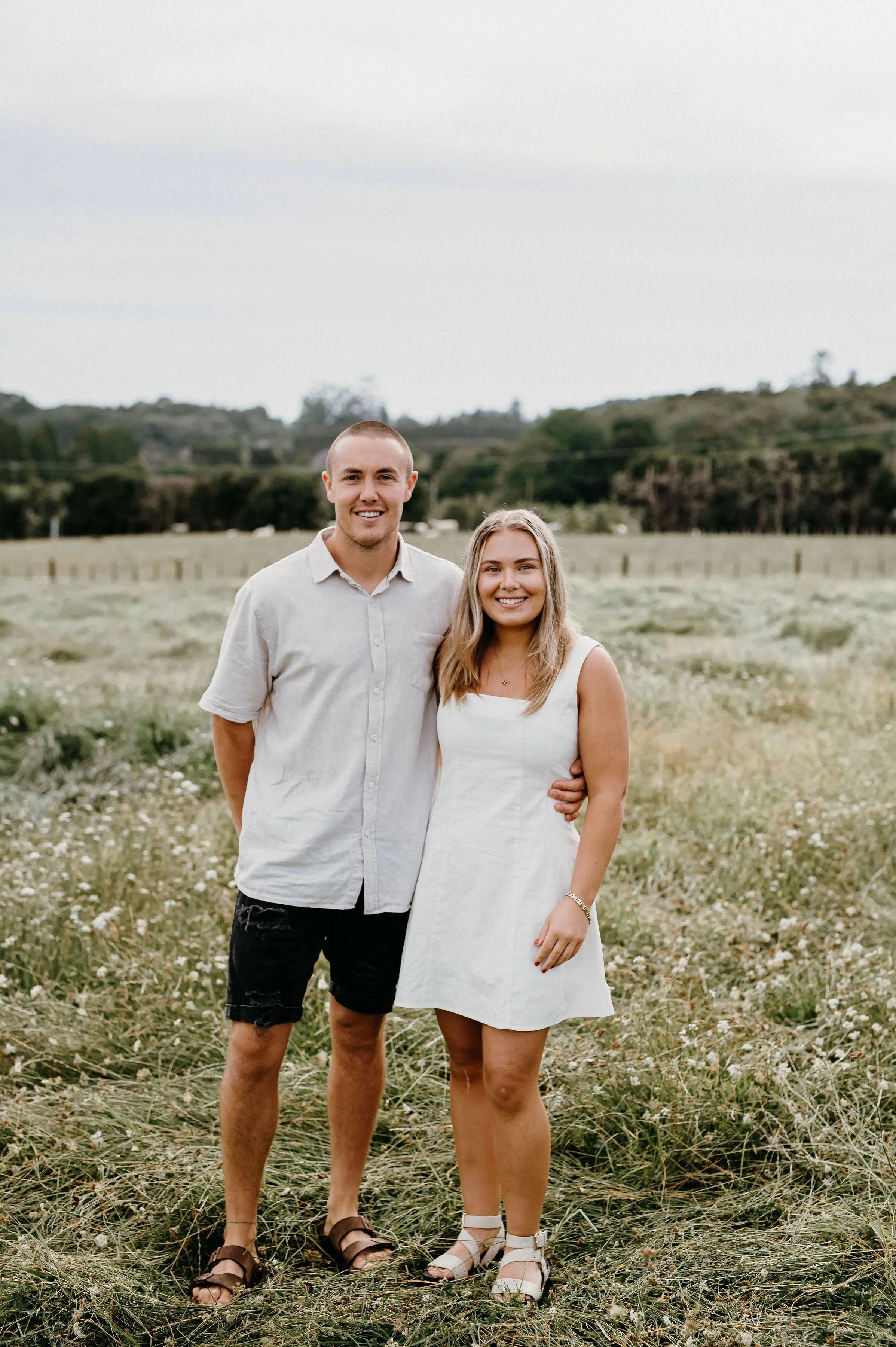 A young man and woman stand together outdoors in a grassy field with rolling hills and trees in the background, smiling at the camera.